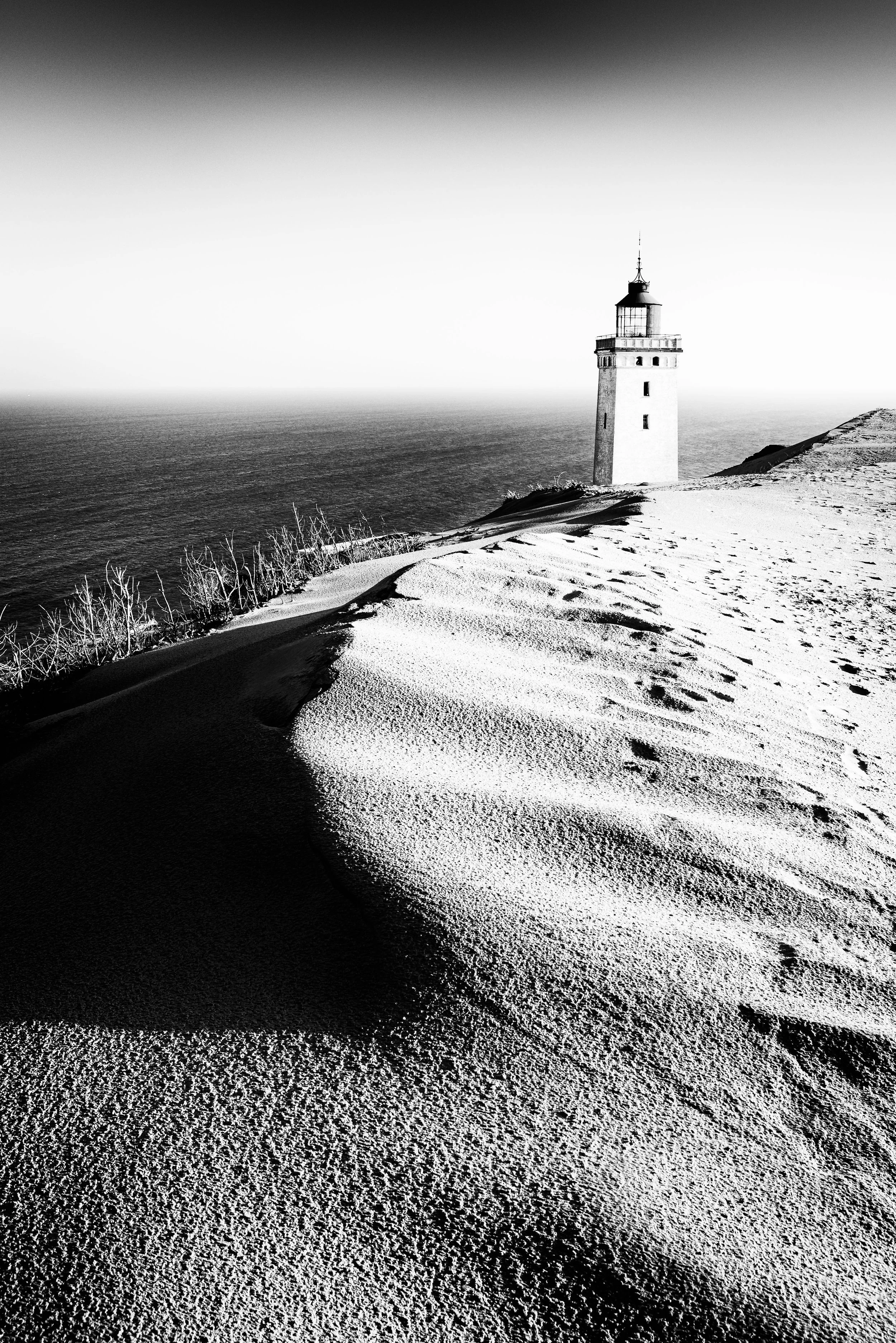 Black and white photo of a lighthouse on a sandy hill with footprints, overlooking the ocean.