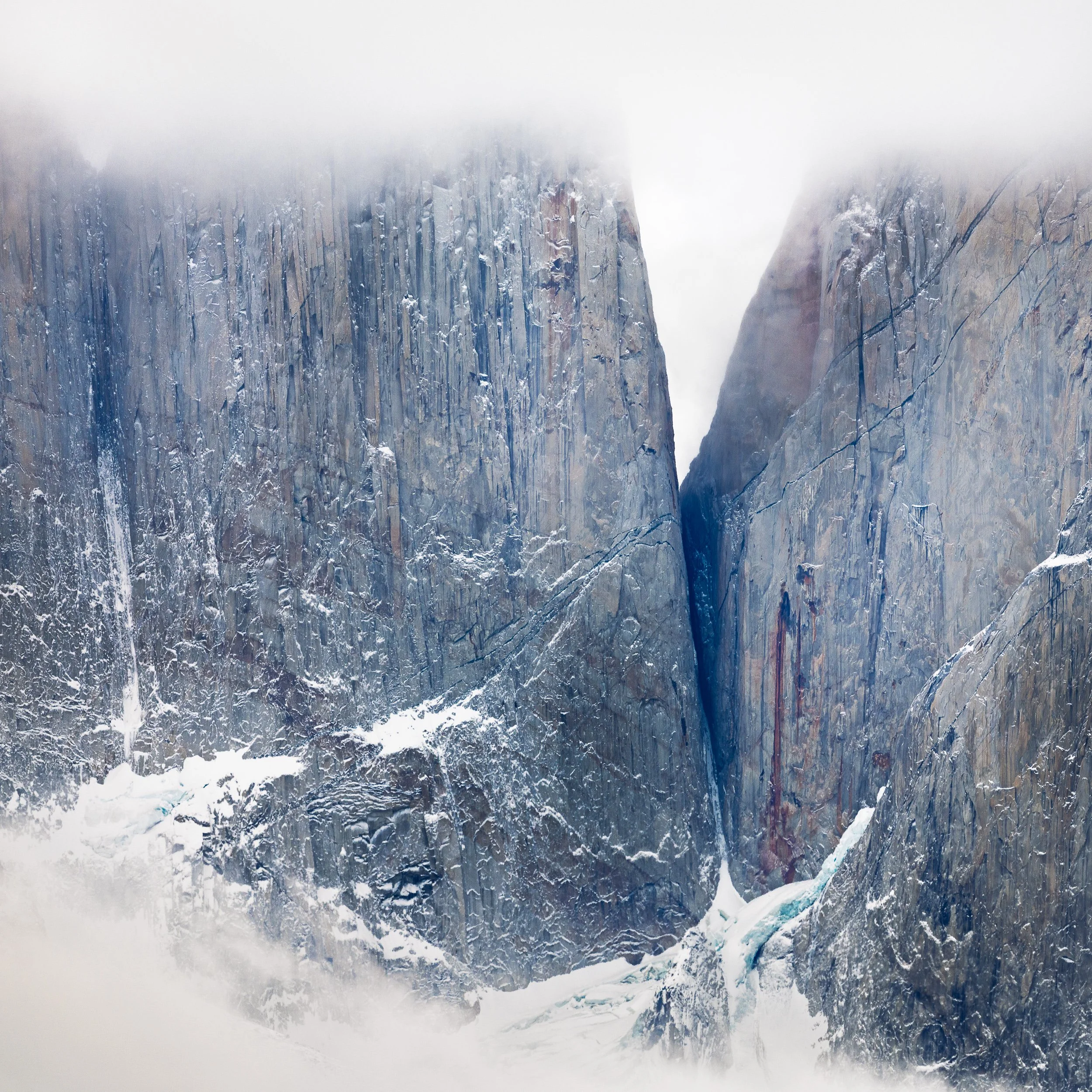 A photograph of steep rocky mountains partially covered in snow and fog, with a narrow icy gorge at the bottom.