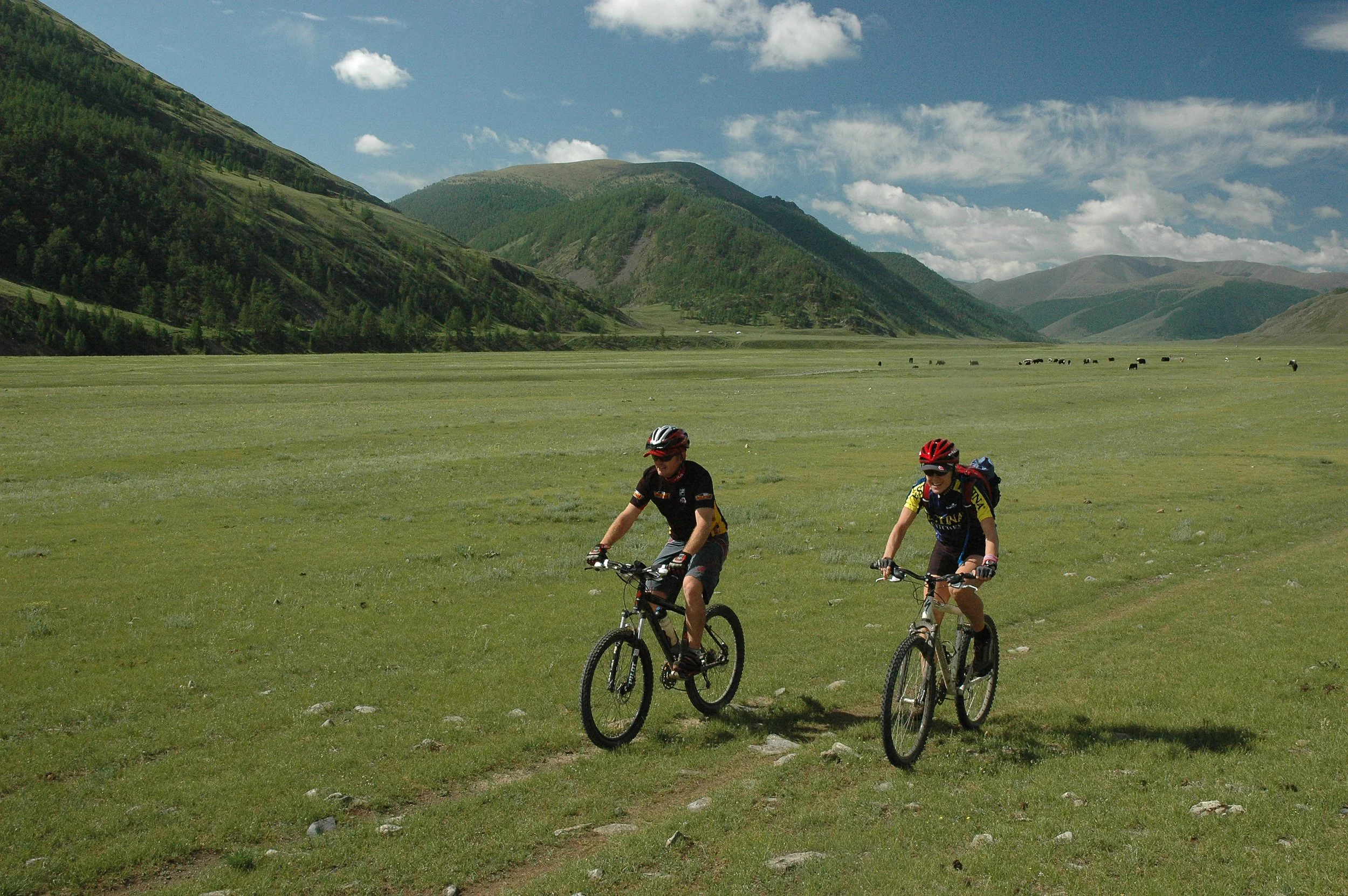 Two children riding mountain bikes on a dirt path in a lush green valley with mountains in the background and a clear blue sky.