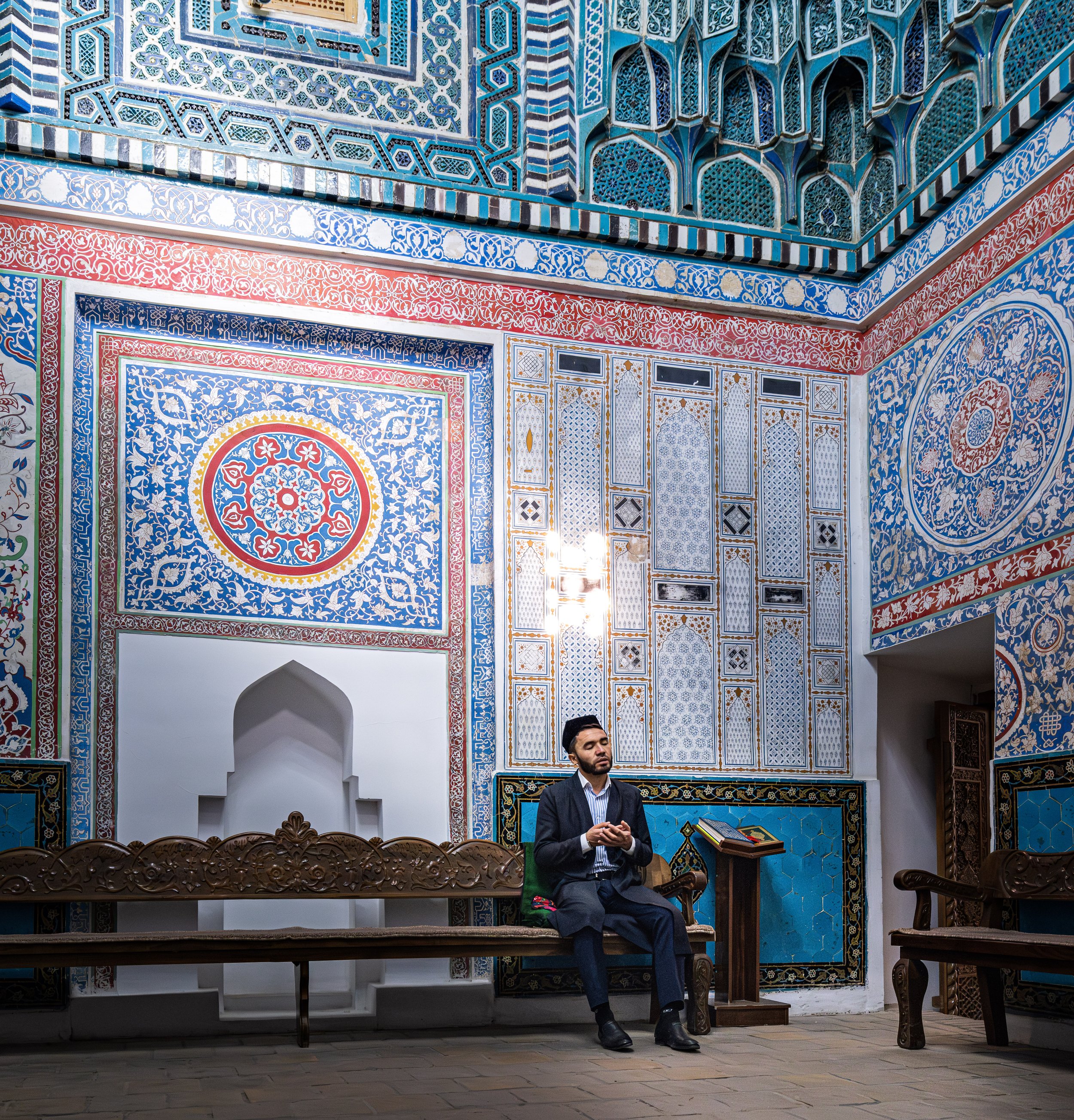 A man sitting on a carved wooden bench inside an ornately decorated room with vibrant blue, red, and gold patterned walls and ceiling, typical of Islamic architecture.