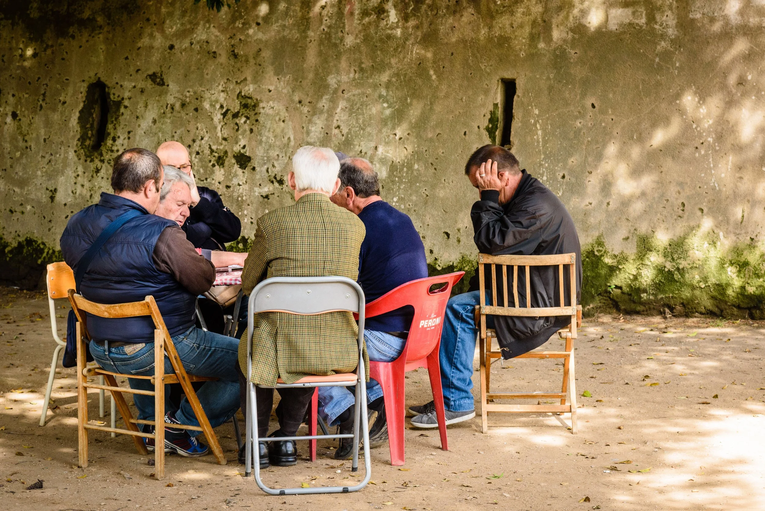 A group of seven men sitting around a table outdoors, some with heads bowed and one appearing distressed, against a moss-covered wall.