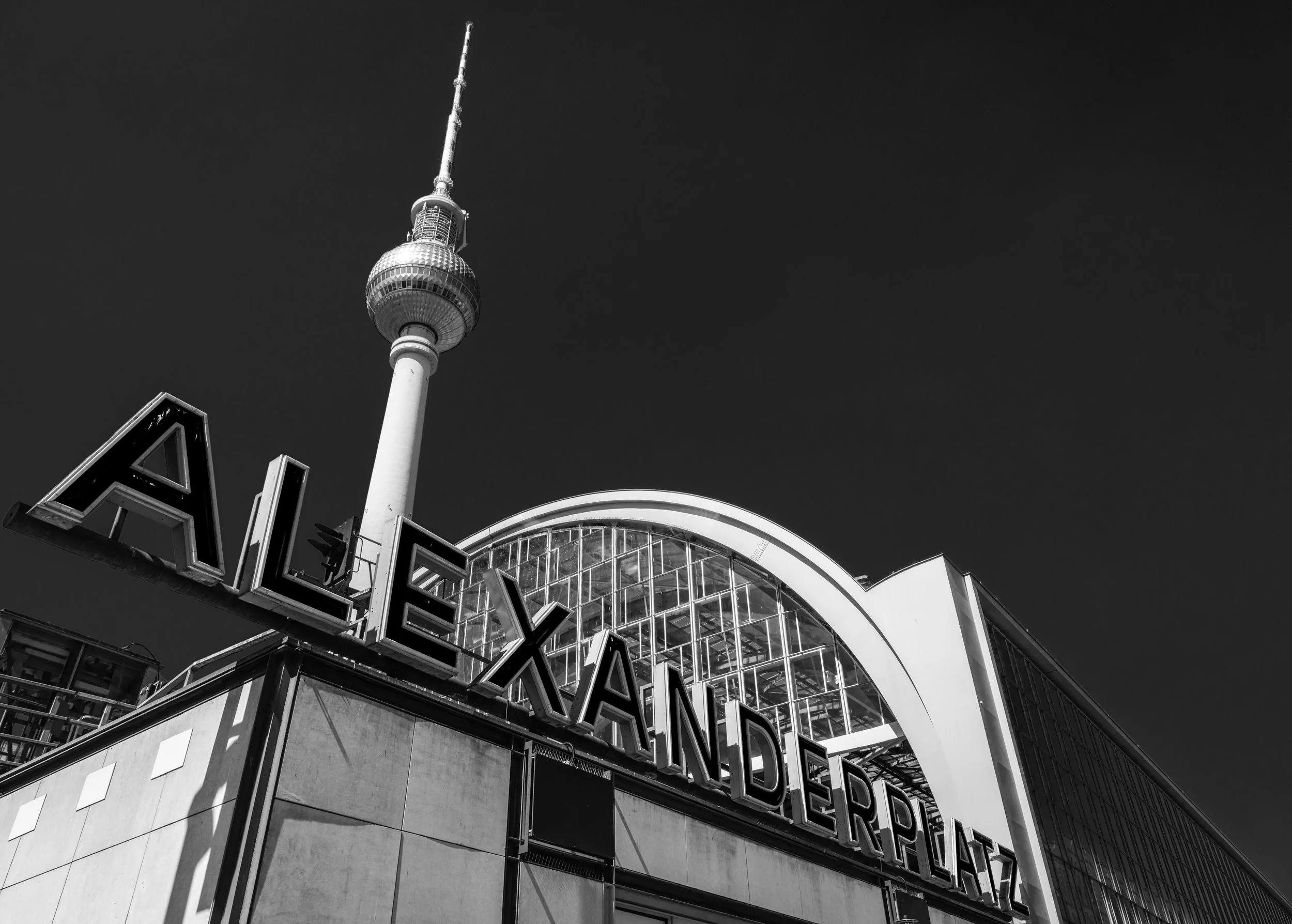 Black and white photo of the Alexanderplatz station in Berlin with a prominent sign and the Fernsehturm tower in the background.