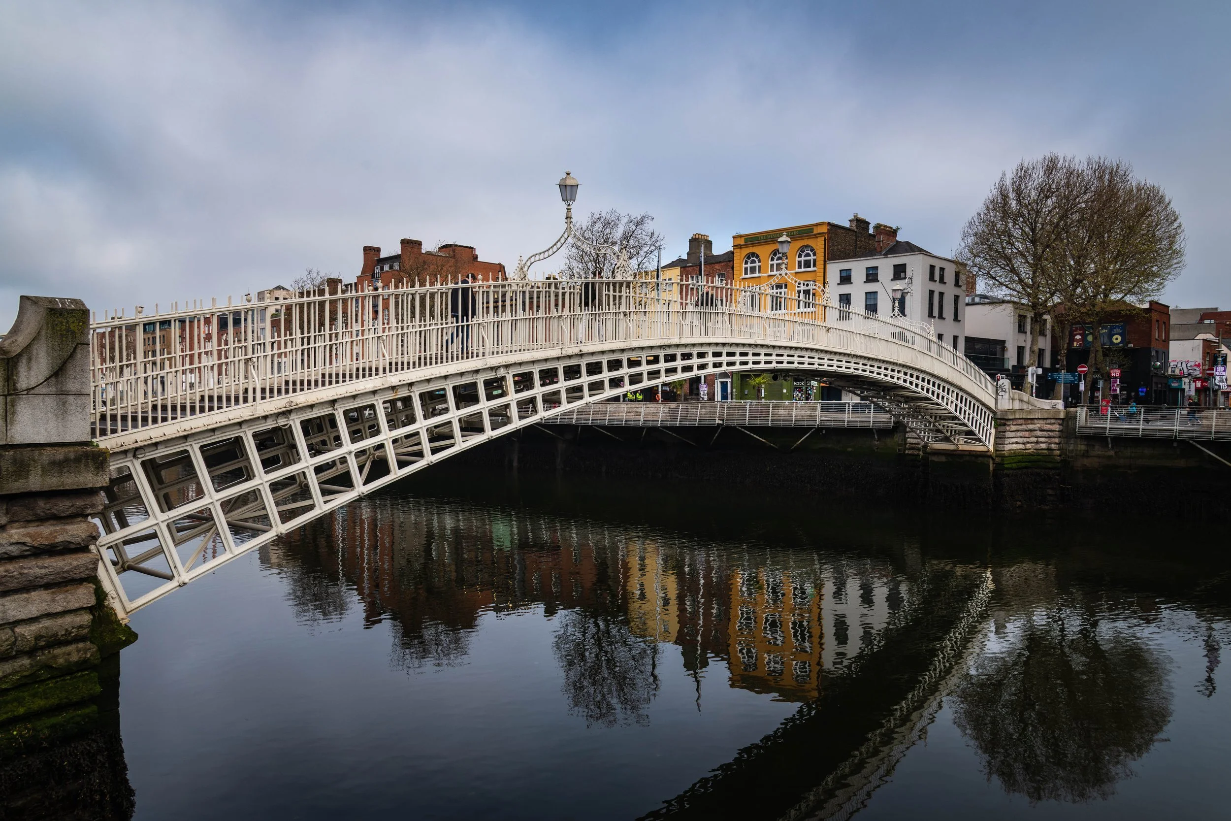 A white arched pedestrian bridge over a river, with buildings and trees in the background on a cloudy day.