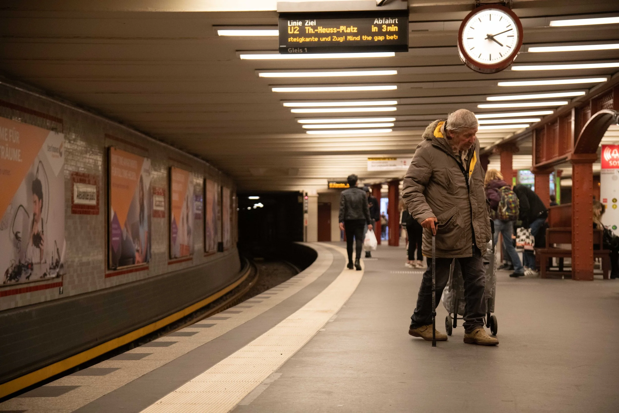 Older man with gray hair, in a brown jacket, using a cane and a walker, waiting on a subway platform.