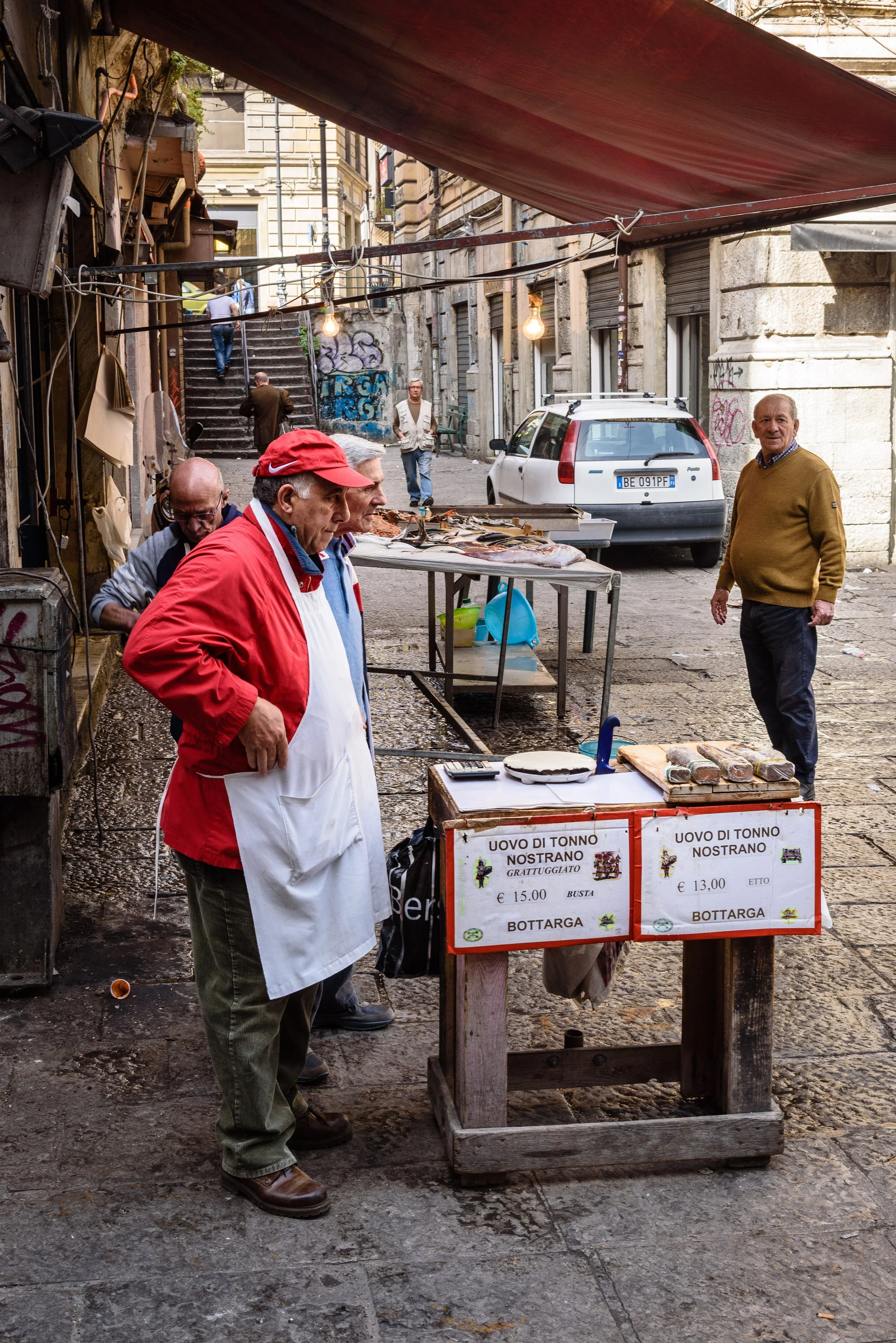 Street market stall selling canned tuna eggs, with three men looking at the display and one man behind the stall in an urban setting.
