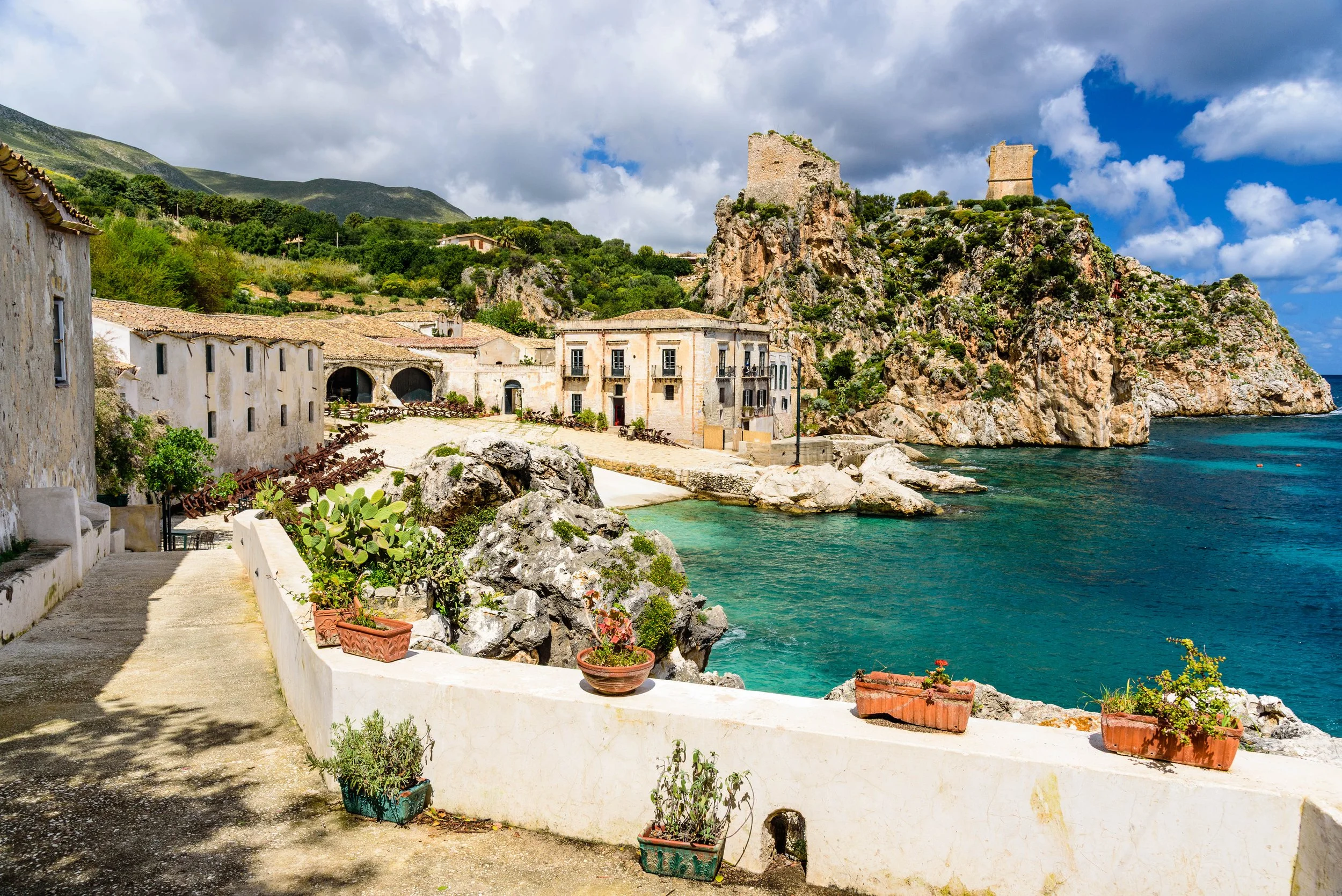 Coastal village with old stone buildings, cacti in pots, rocky cliffs, and a castle ruins on a hill under a partly cloudy sky, overlooking a turquoise sea.