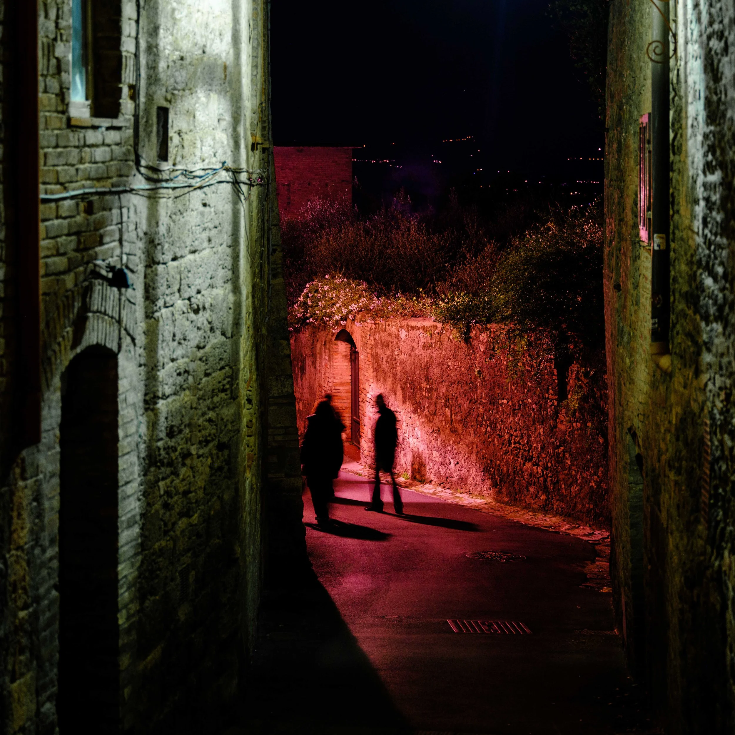 Nighttime alleyway with two silhouetted people walking, illuminated by pinkish streetlights, flanked by old stone buildings, with a dark sky in the background.
