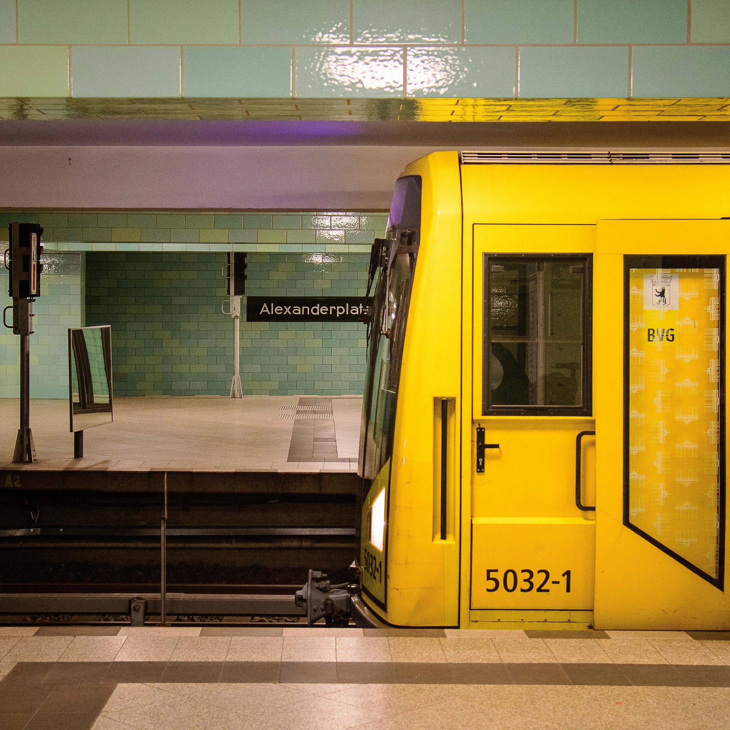 A yellow train at the Alexanderplatz subway station in Berlin, Germany, with a green tiled wall and platform in the background.