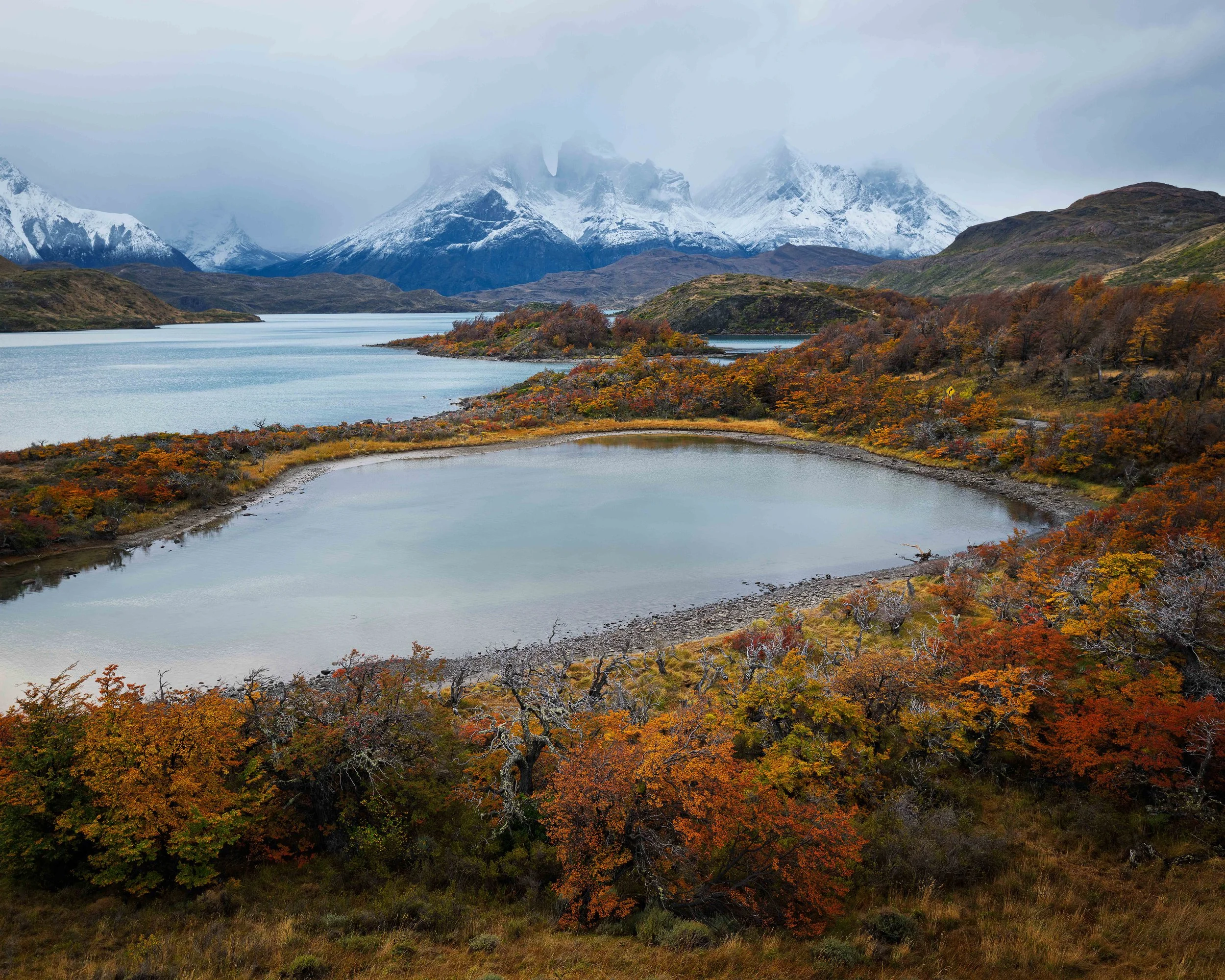 A landscape featuring a lake with autumn-colored trees, mountains in the background with snow, and cloudy skies.