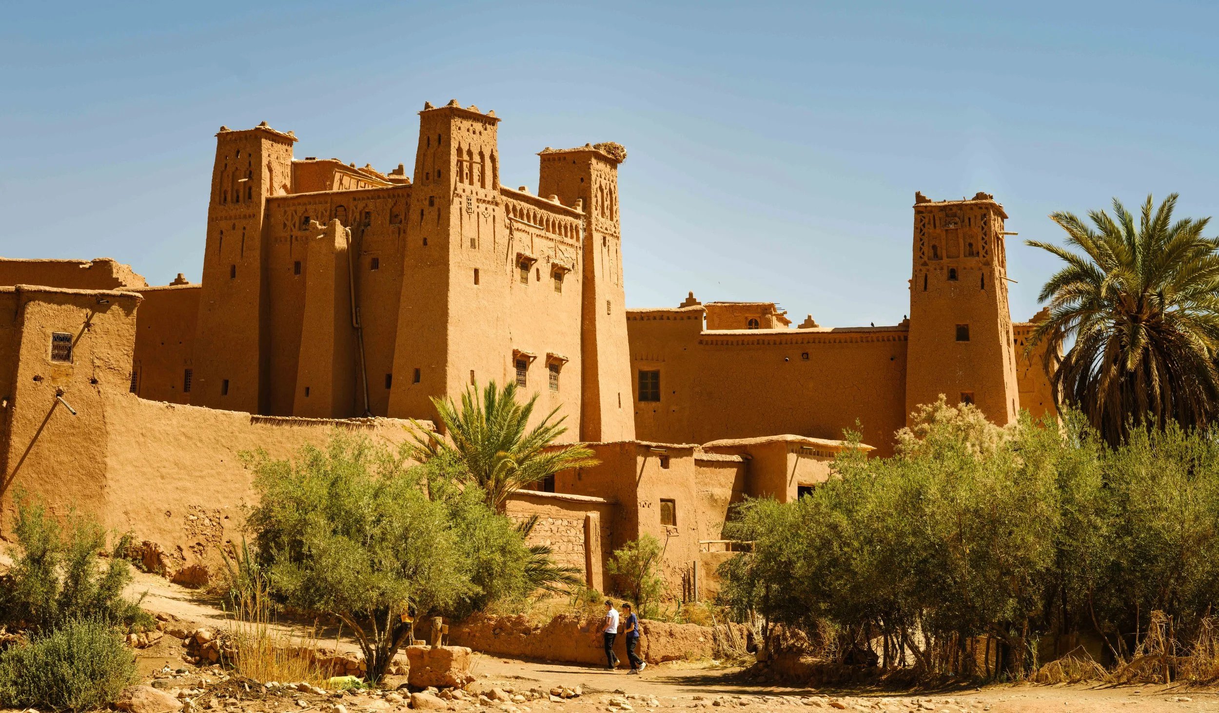 A large traditional desert fortress made of mud brick with tall towers, surrounded by palm trees and desert vegetation under a clear blue sky.