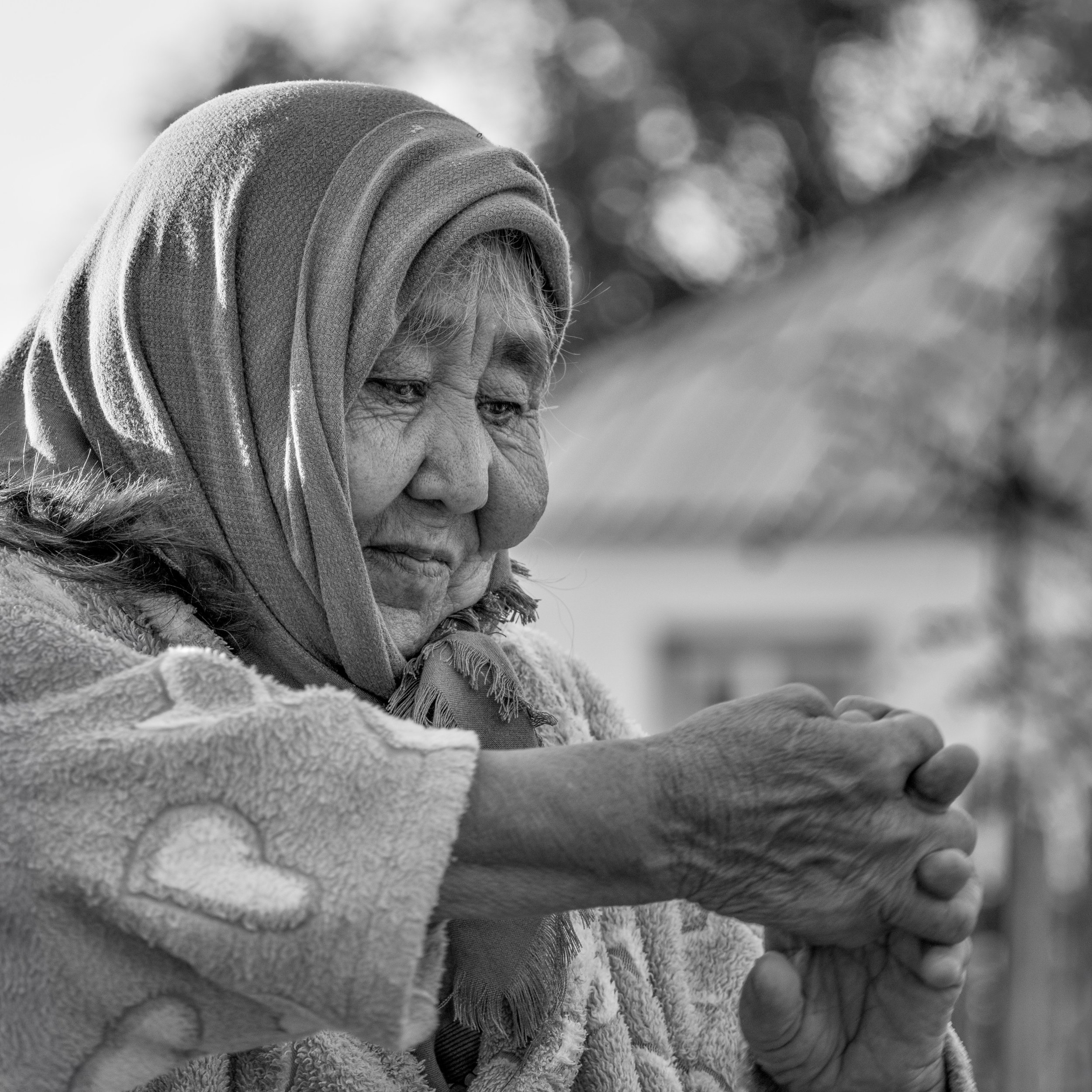 Black and white photo of an elderly woman wearing a headscarf praying with clasped hands outside.