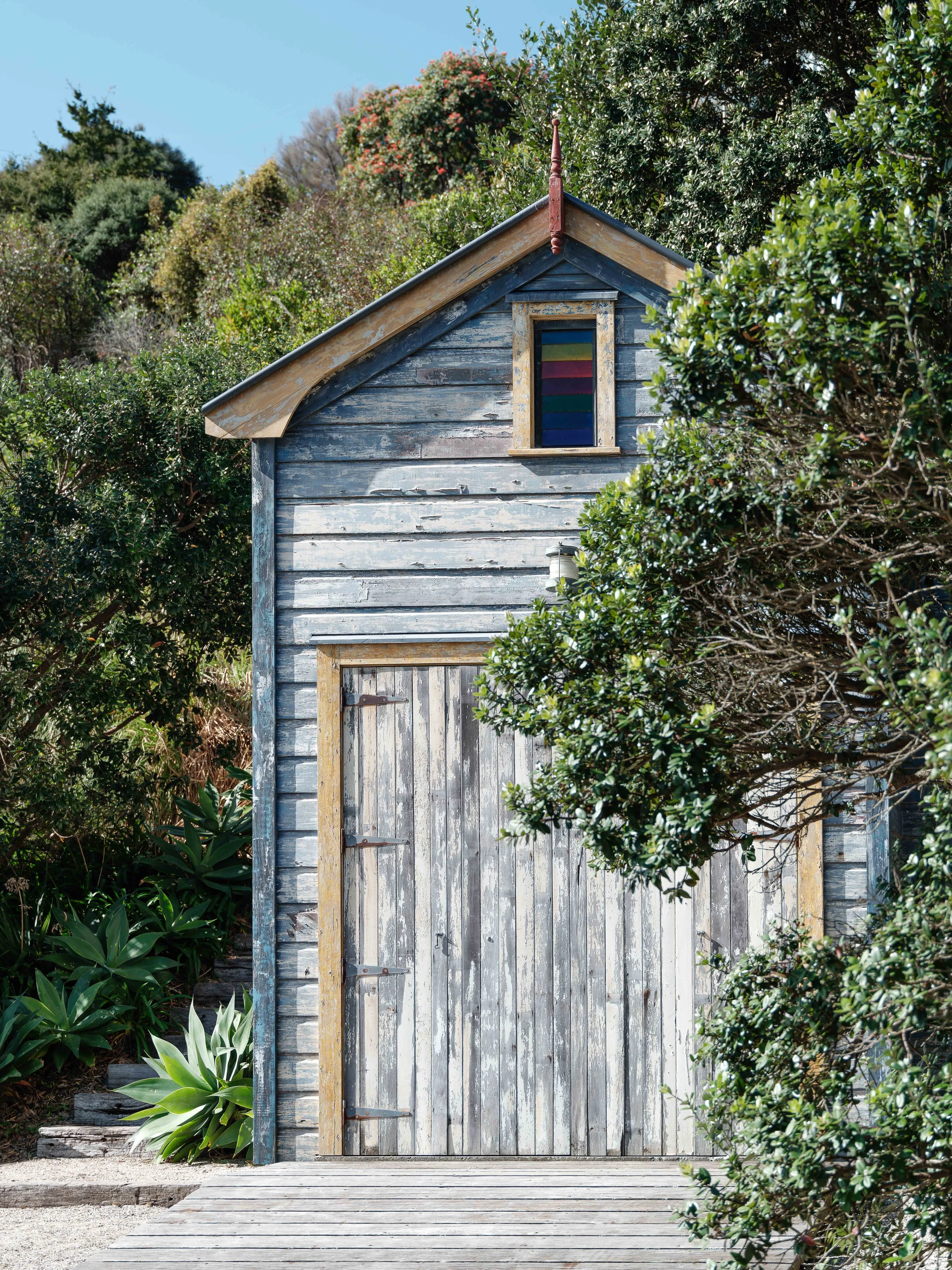 A weathered wooden shed with a double door, surrounded by green shrubs and trees, with a small window near the roof and a blue sky above.