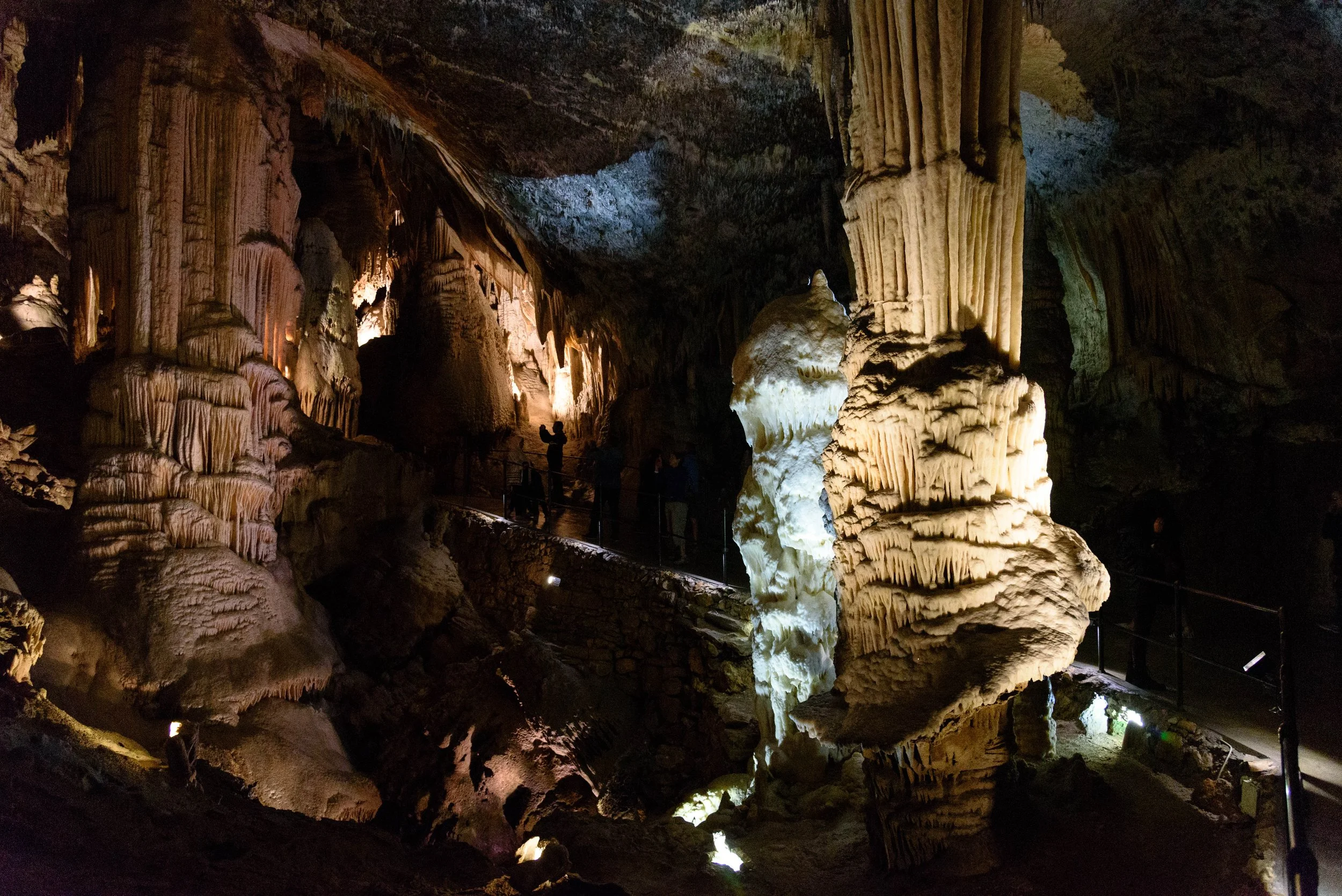 People walking inside a dark cave with large, illuminated stalagmites and stalactites.