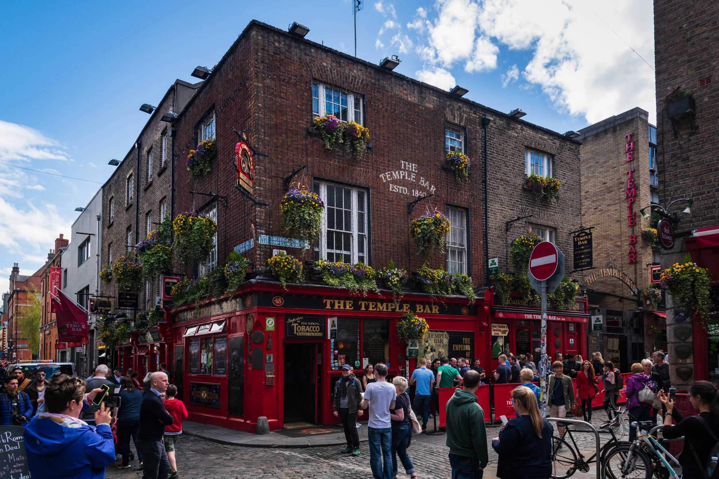 Crowds gather outside The Temple Bar, a red building with hanging flower baskets on a cobblestone street in Dublin, Ireland.