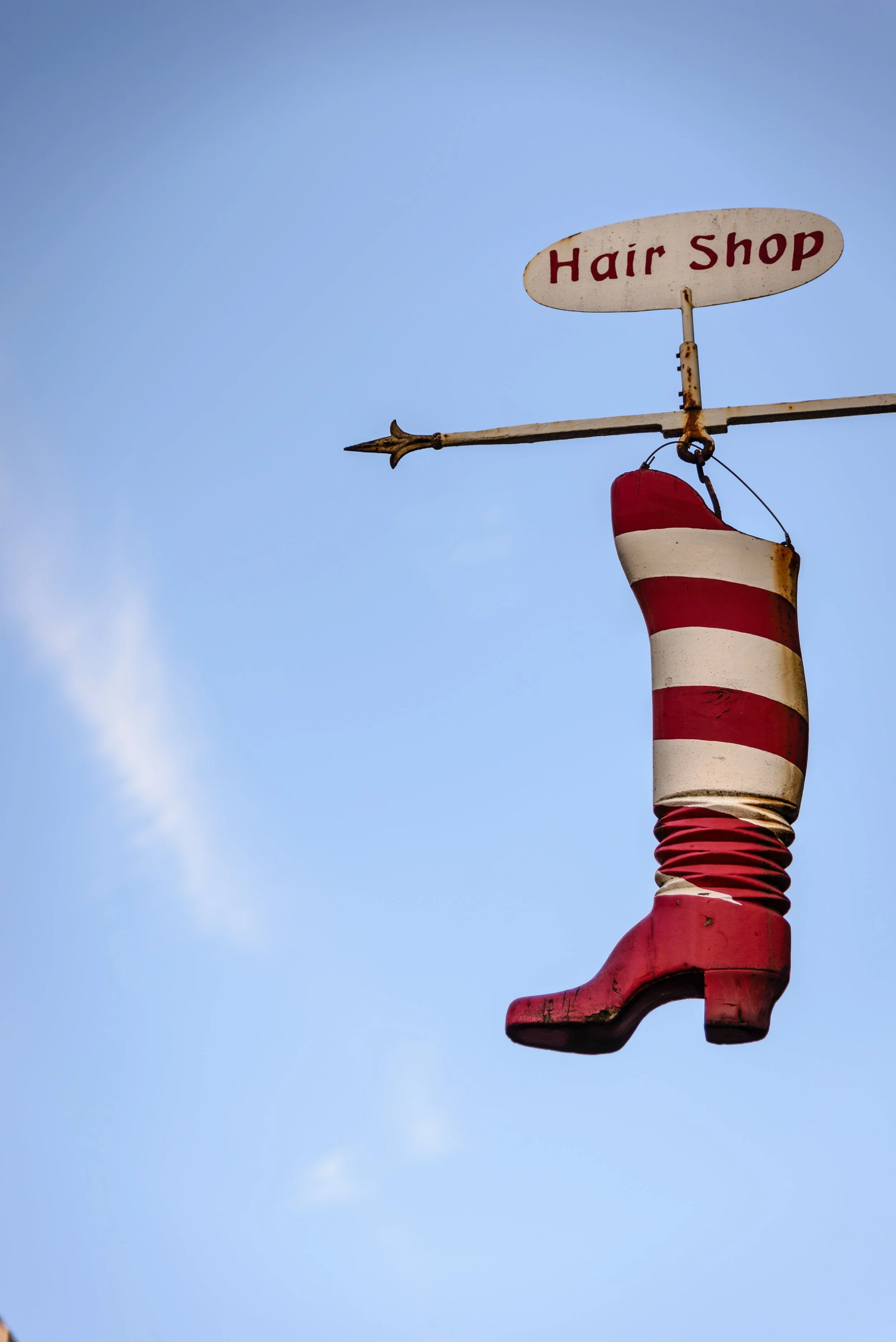 A weathered sign for a hair shop featuring a red and white striped boot-shaped hanging sign against a clear blue sky.