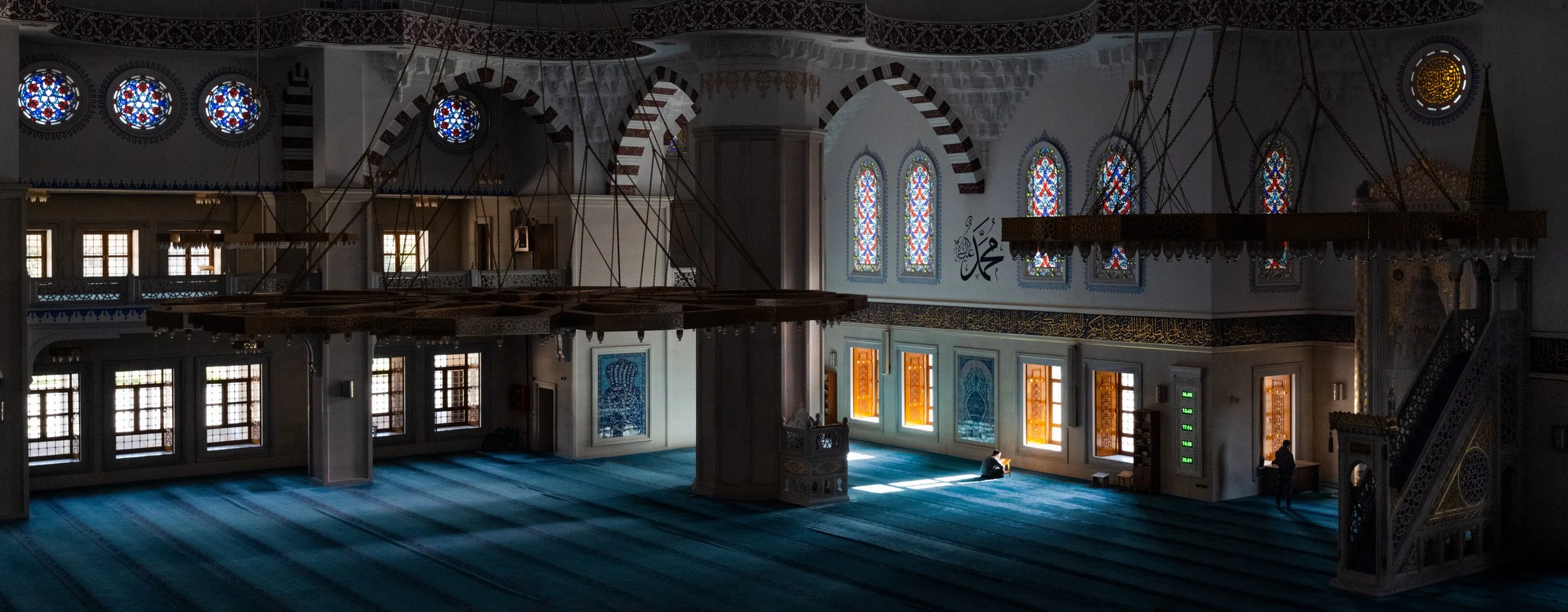 Interior of a mosque with stained glass windows, blue carpet, and a few people sitting and standing.