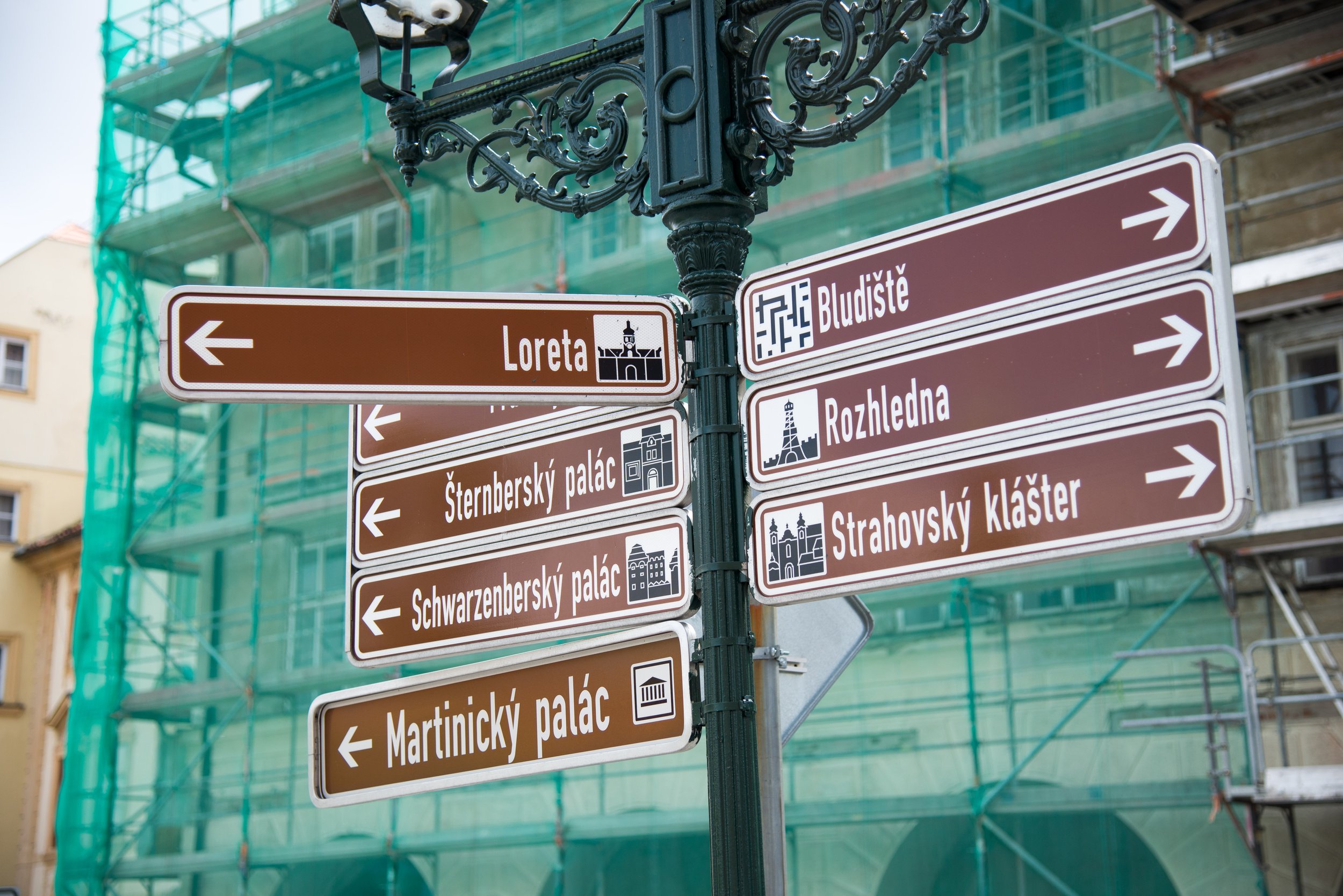 Street signpost with directions to various historic sites and landmarks, including Loreta, Bludiště, Šternberský palace, Rozhledna, Schwarzenberský palace, Strahovský klášter, and Martinic palace, in a city with building scaffolding in the background