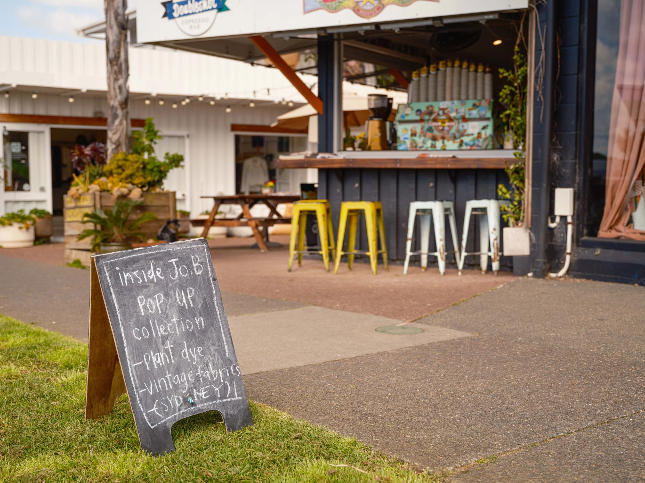 A black chalkboard sign outside a shop with handwritten white chalk text advertising a pop-up collection featuring plant dye, vintage fabrics, and an event. In the background, there are three yellow and three white metal stools at a wooden bar, a pic