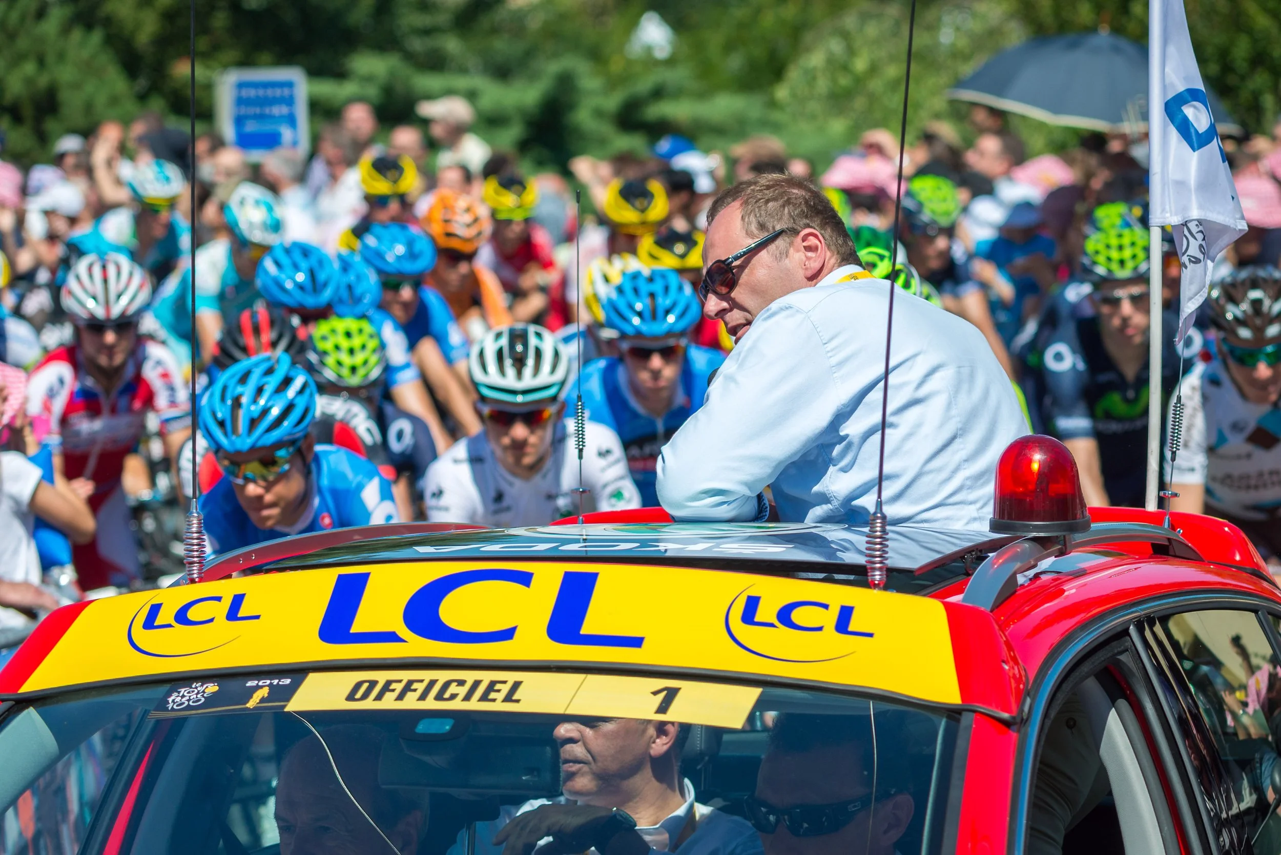 A man in a white shirt sitting on a red official car with yellow and blue liveries, speaking to a large group of cyclists wearing helmets, at a cycling event.