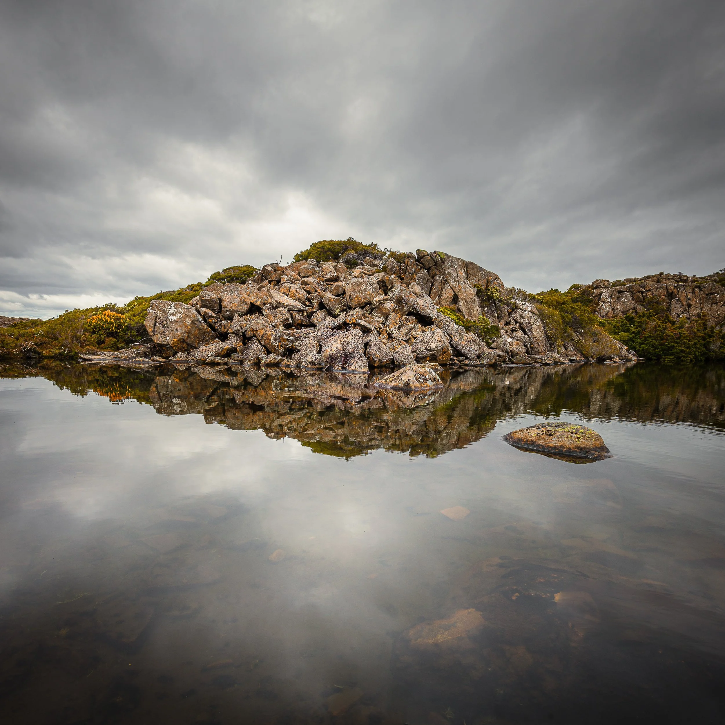 A rocky island reflected in calm water under a cloudy sky.