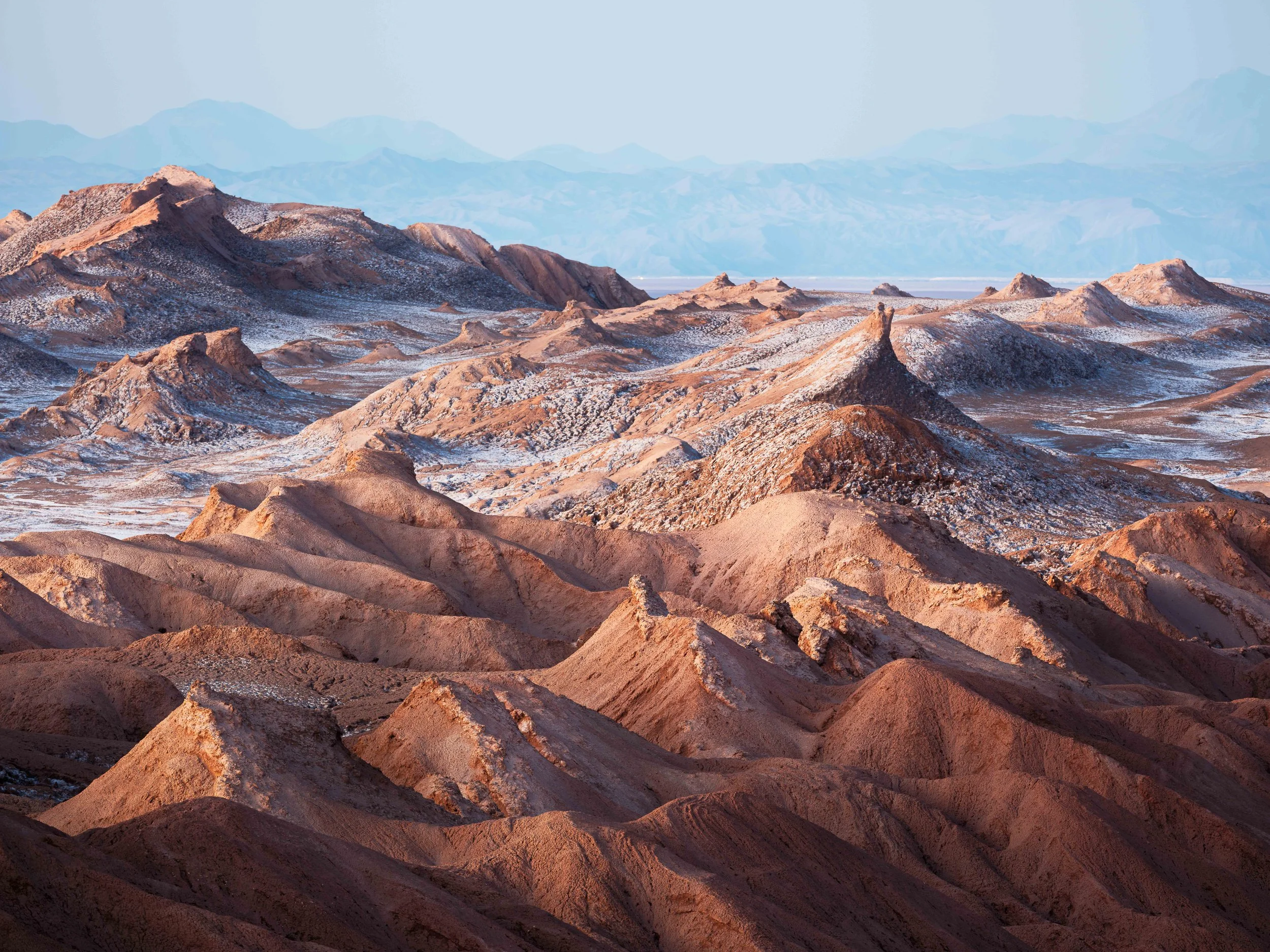 Mountain range with reddish-brown and snow-capped peaks, desert-like terrain, and distant blue mountains under a clear sky.