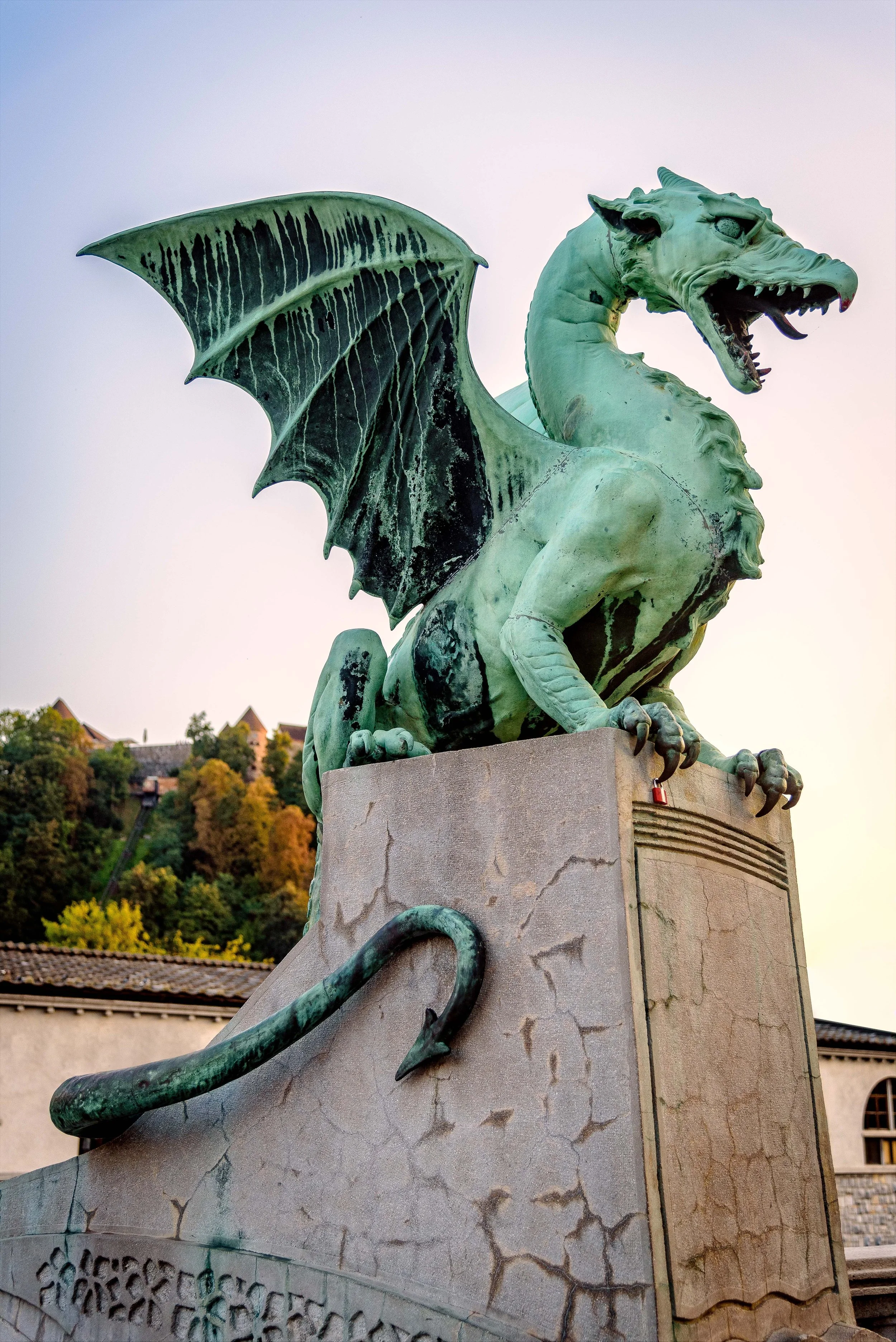 A large bronze statue of a dragon with wings, sitting on a stone pedestal, surrounded by trees and buildings in the background.