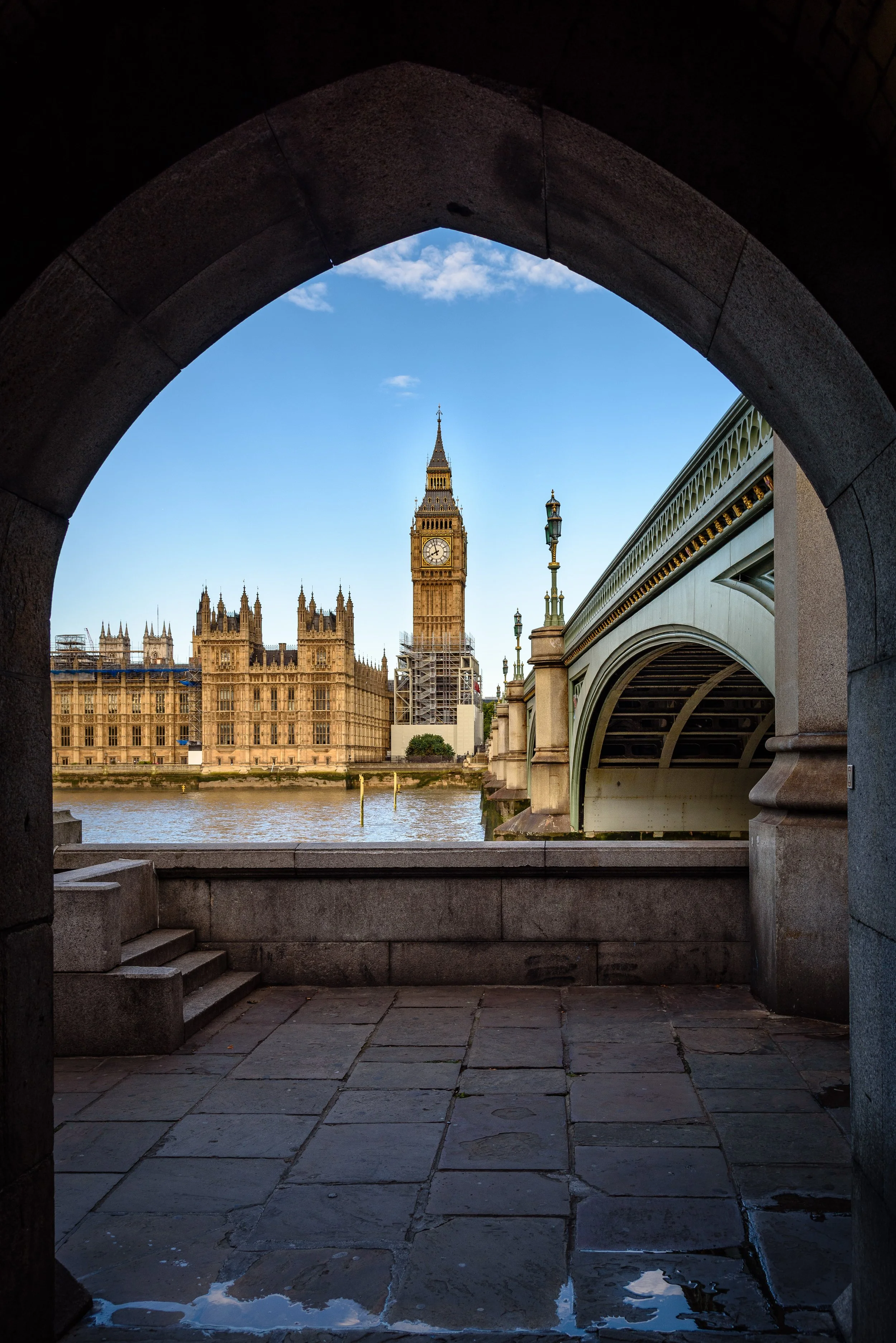 View through a stone archway showing Big Ben and the Houses of Parliament in London, England, with Westminster Bridge to the right and a clear blue sky.