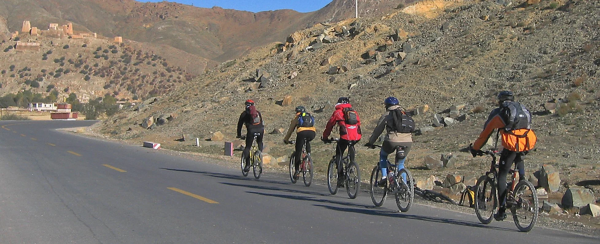 Five cyclists riding along a mountain road with rocky terrain and hills in the background.