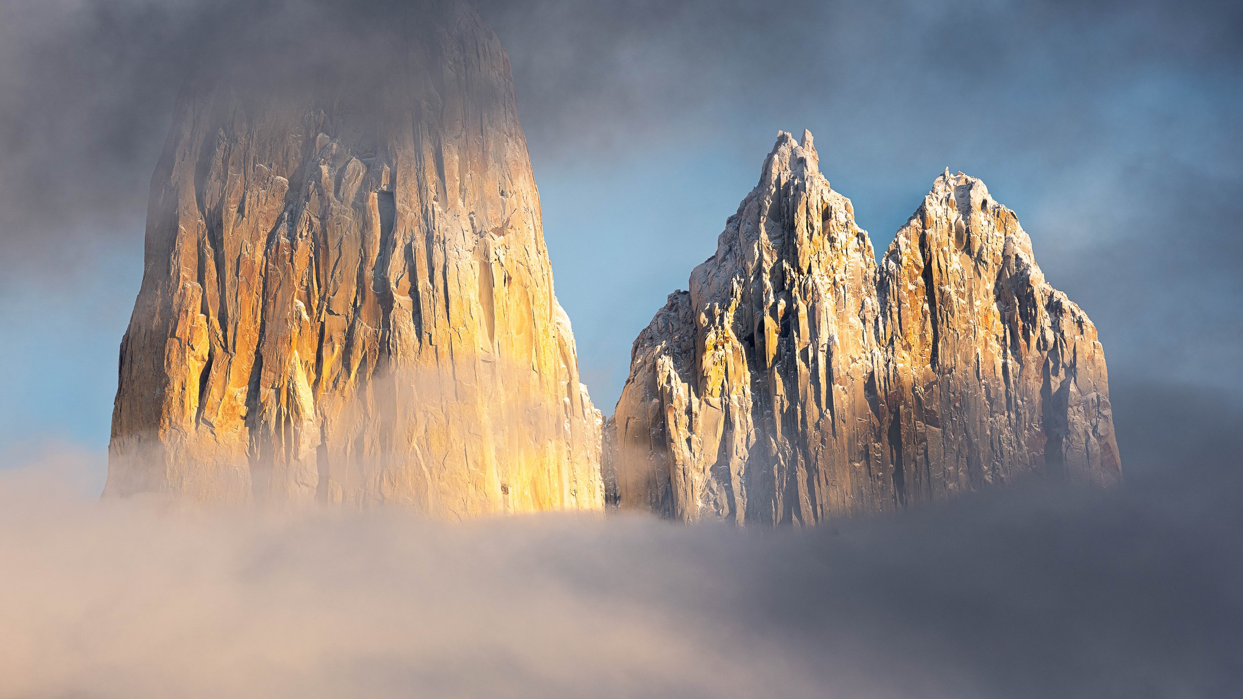 Three mountain peaks surrounded by clouds and fog, with sunlight illuminating the rocky surface.