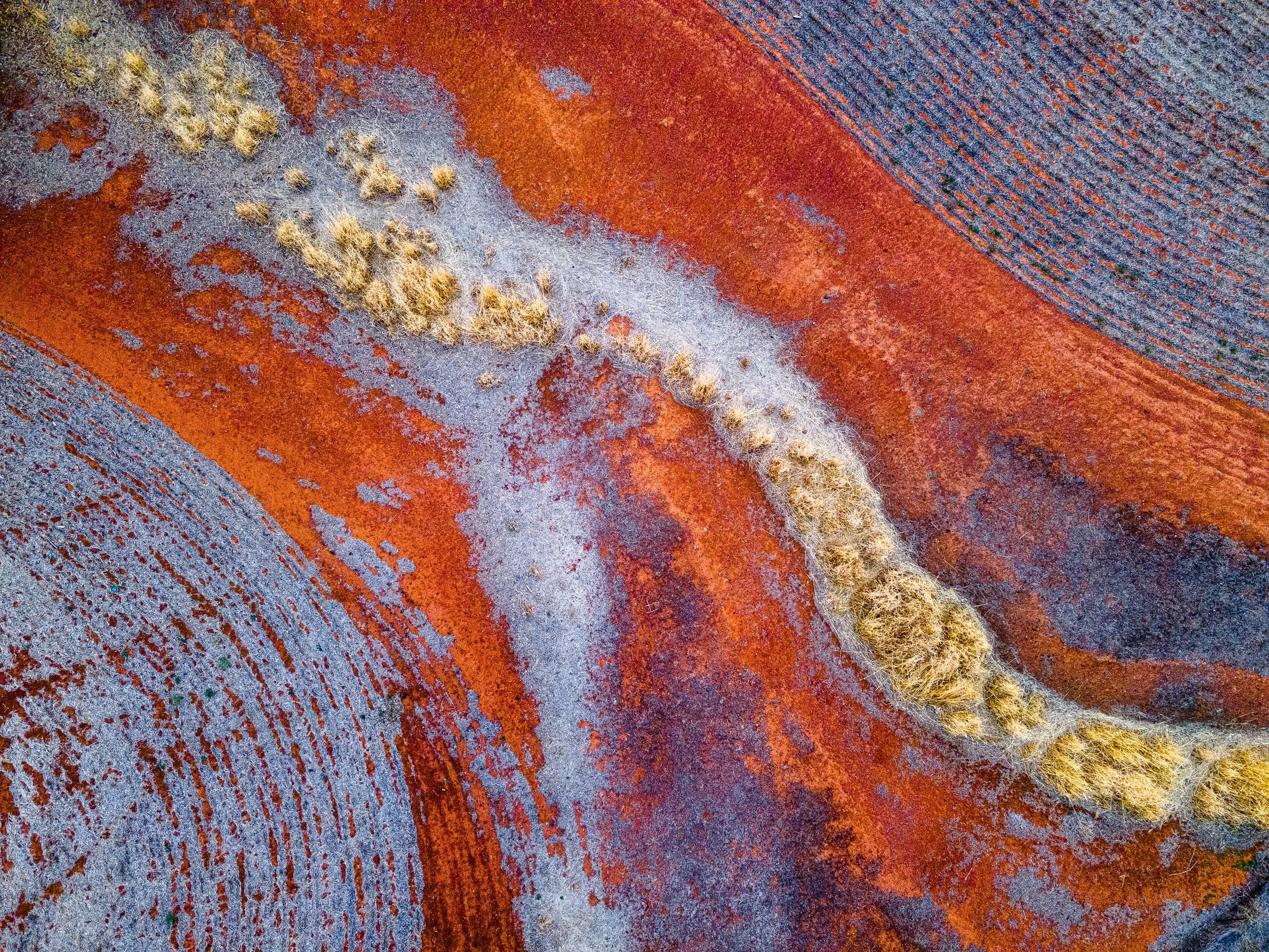 A close-up aerial view of various colorful mineral deposits with orange, blue, and gray hues and a line of fuzzy, yellowish moss or lichen running diagonally through the center.