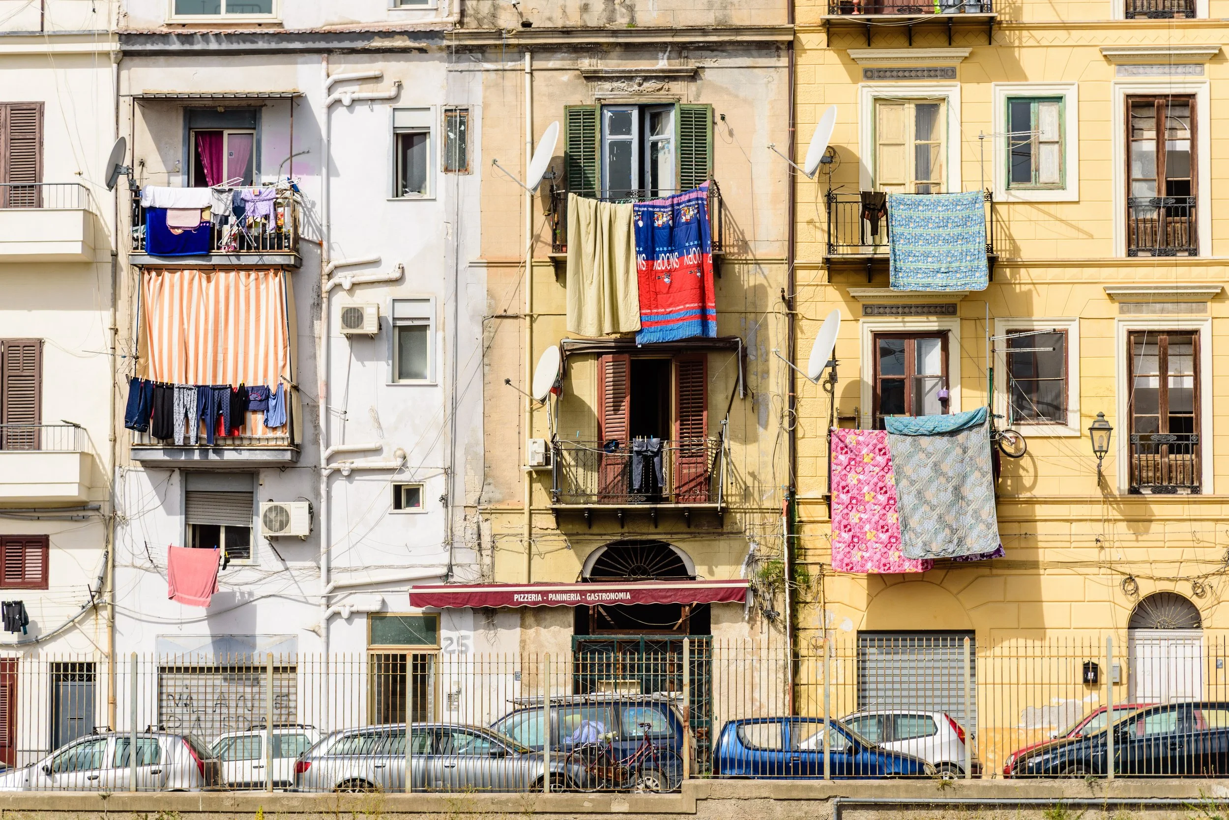 Facade of three colorful European-style apartment buildings with laundry hanging on balconies and windows, parked cars in front, and a restaurant beneath the central building with a red awning.
