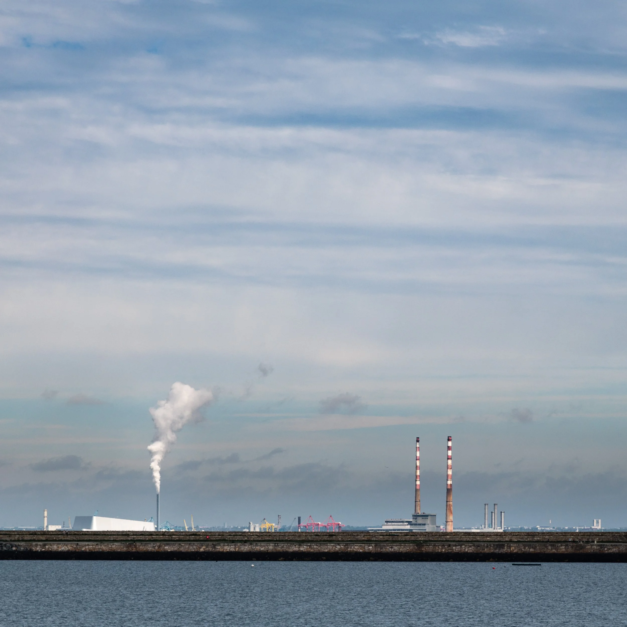 Industrial factory with smokestacks releasing smoke, located near body of water under a cloudy sky.
