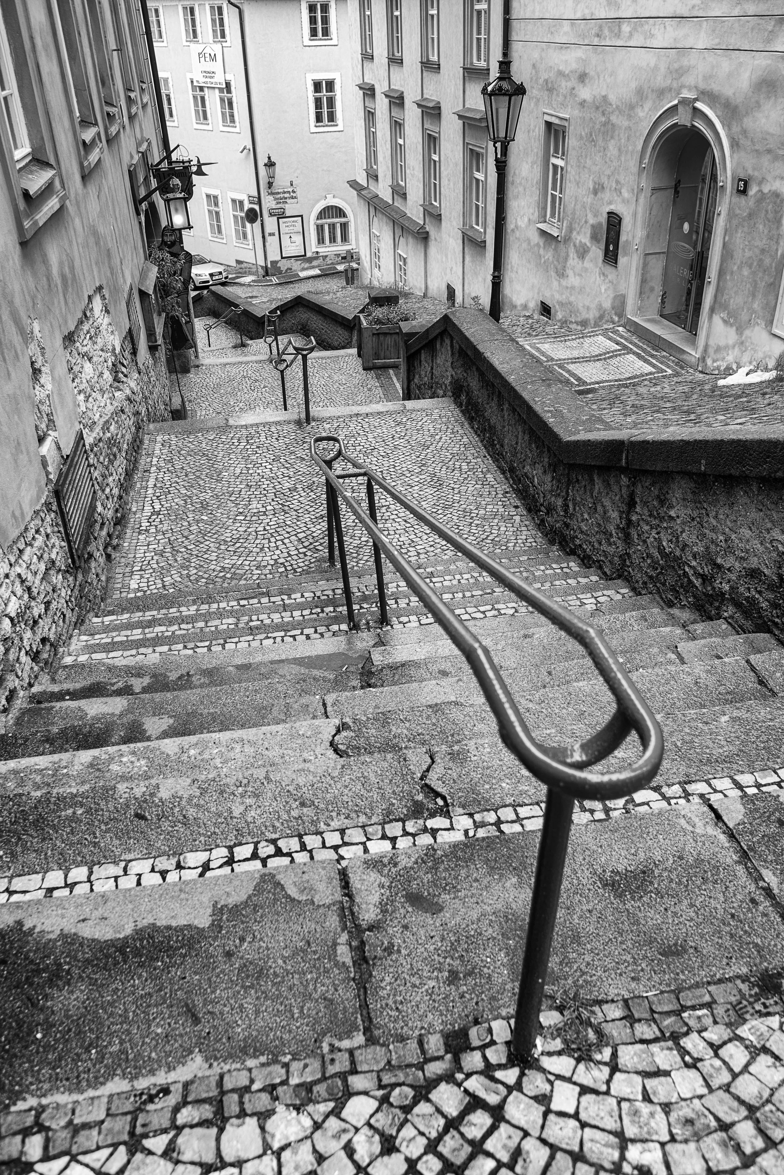 Black and white photo of a steep outdoor stone staircase with metal handrails on both sides, leading down between old buildings with cobblestone pavement at the bottom.