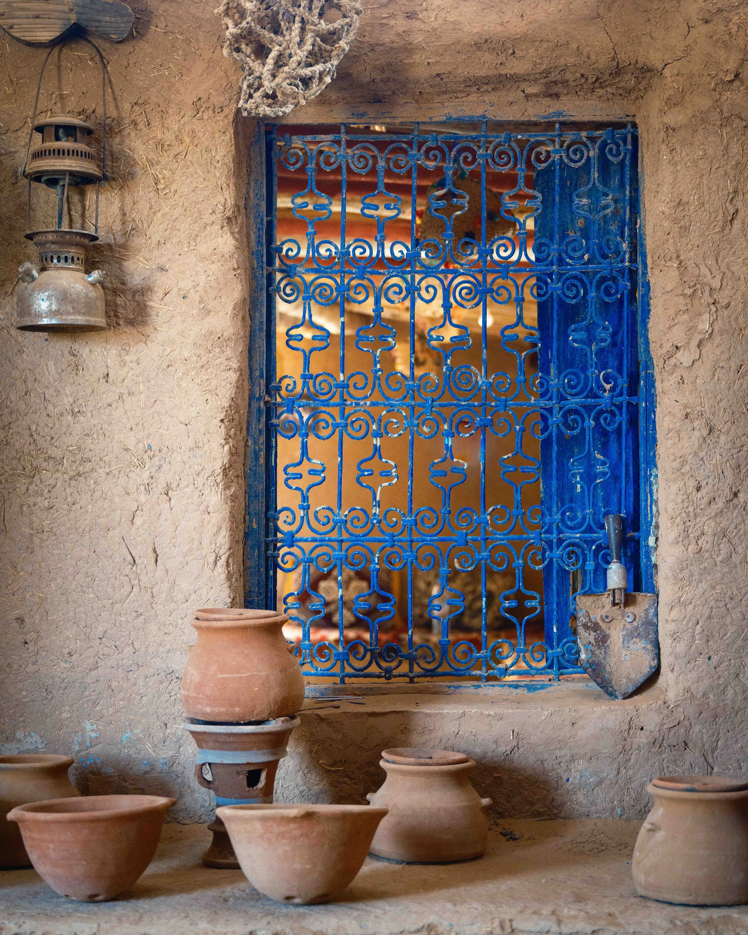 Clay pots placed on the ground in front of a rustic wall with a blue iron grille window, an old shovel leaning against the wall, a hanging lantern, and a rope hanging above.