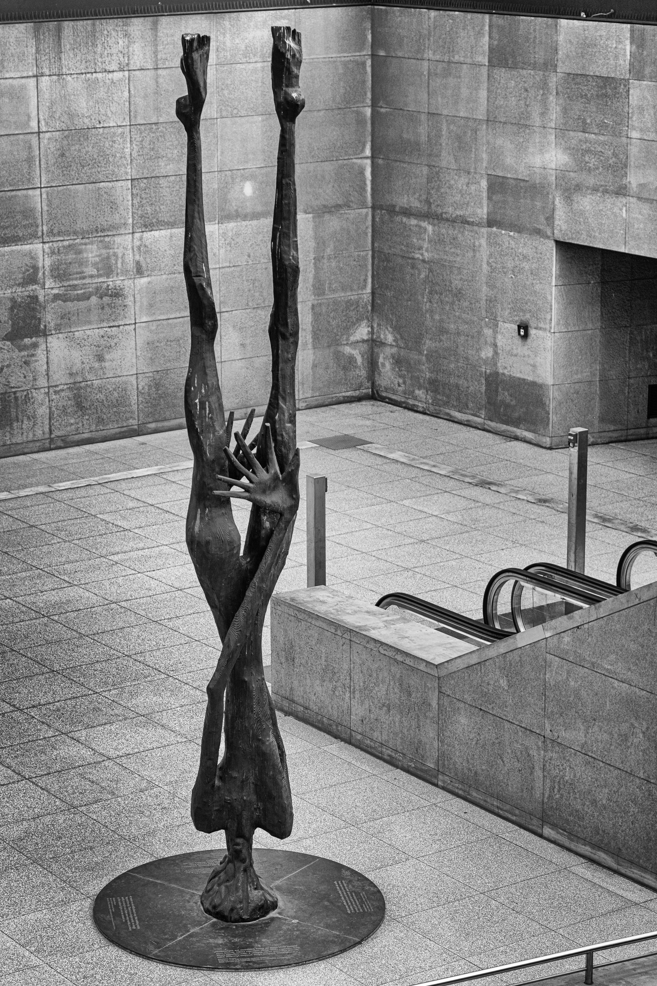 Black and white photo of a modern sculpture of a hand emerging from a human figure, both elongated and abstract, located in a spacious indoor area with tiled floors and stone walls.