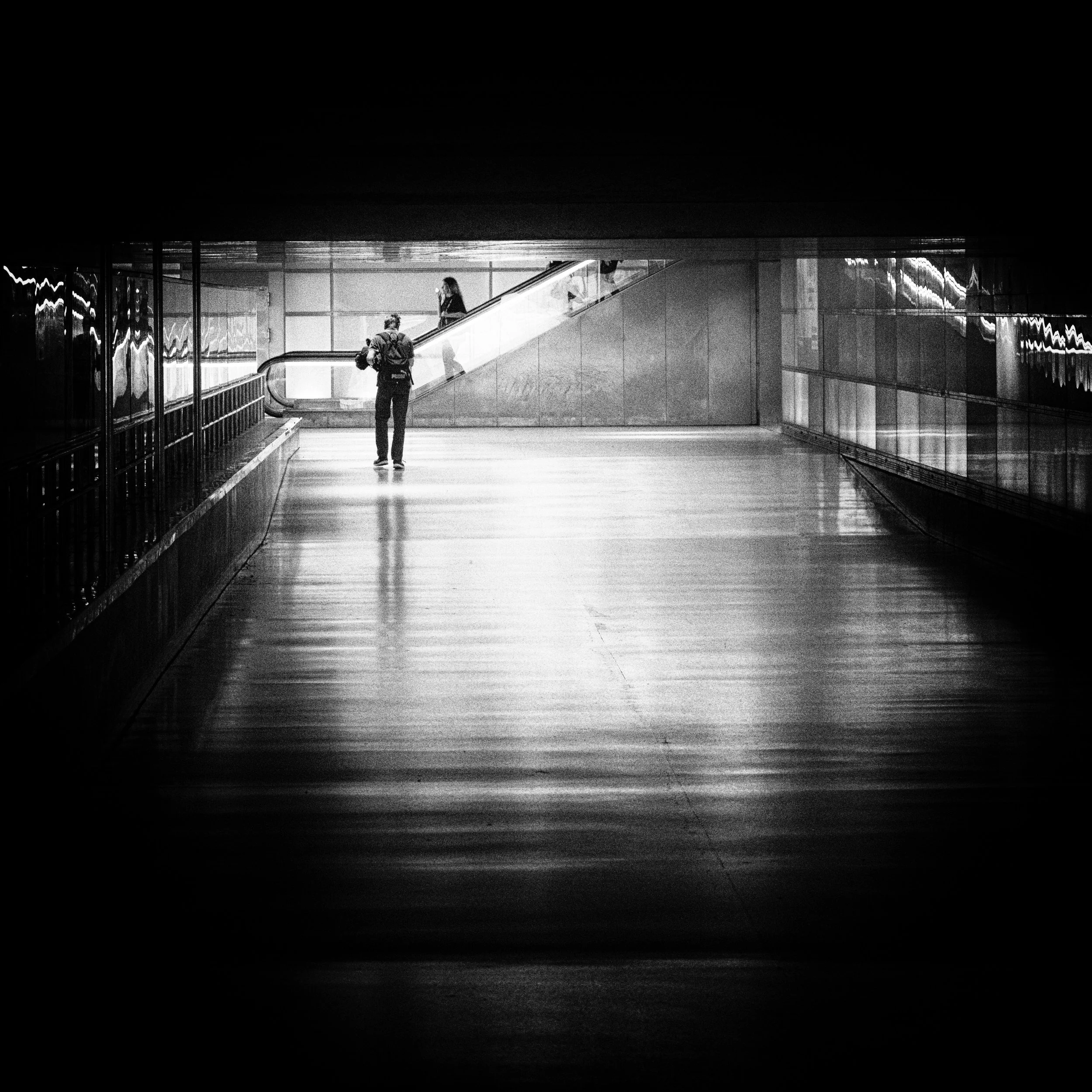 Black and white photograph of a person with a backpack walking in an underground walkway or tunnel, heading toward an escalator. The scene is dimly lit, with reflections on the floor and walls.