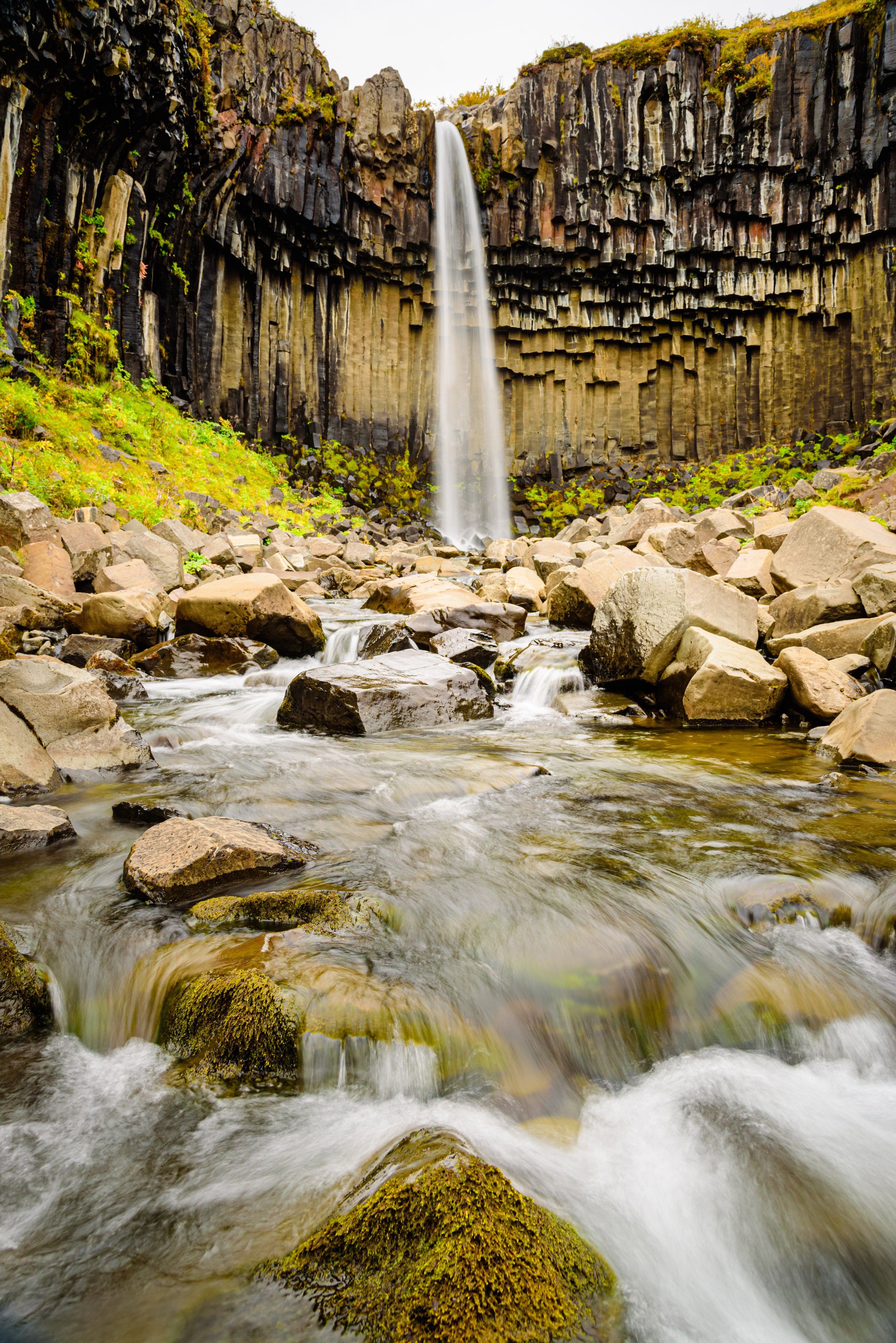A tall waterfall cascading down a basalt cliff into a rocky stream with moss-covered stones.