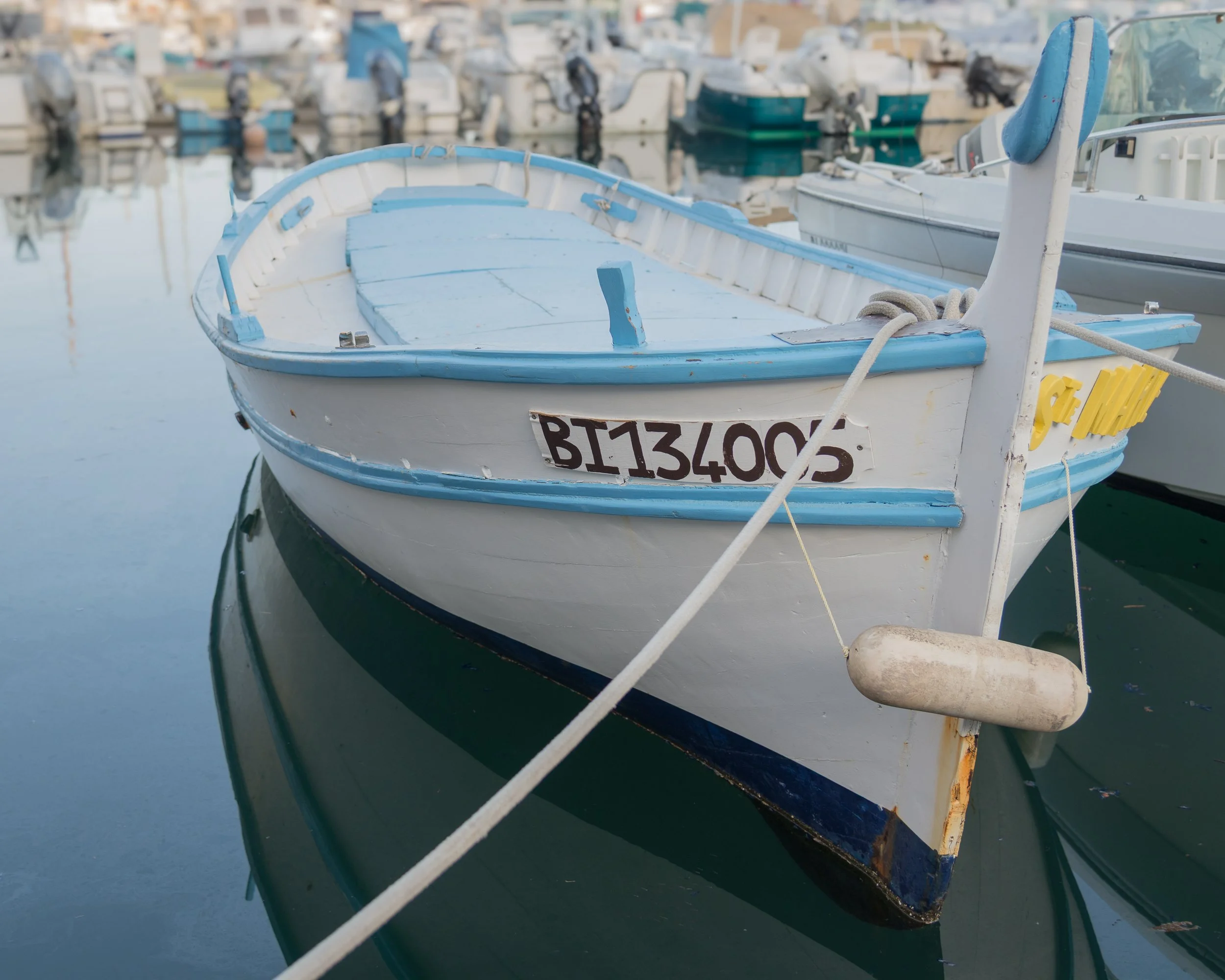 A small white and blue rowboat docked at a marina with other boats in the background, reflected in calm water.