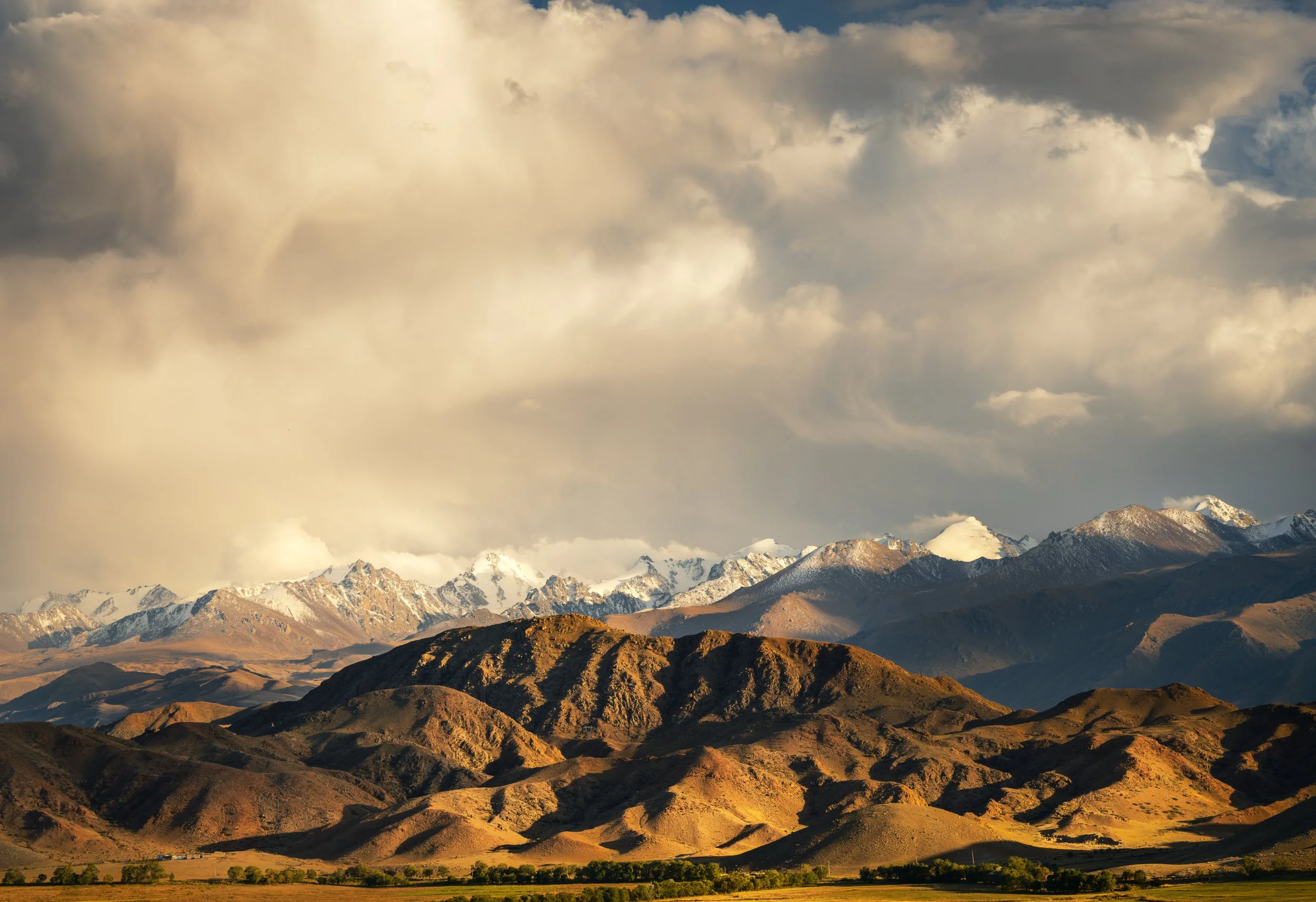 Mountain range with snow-capped peaks under cloudy sky, rolling hills in the foreground.
