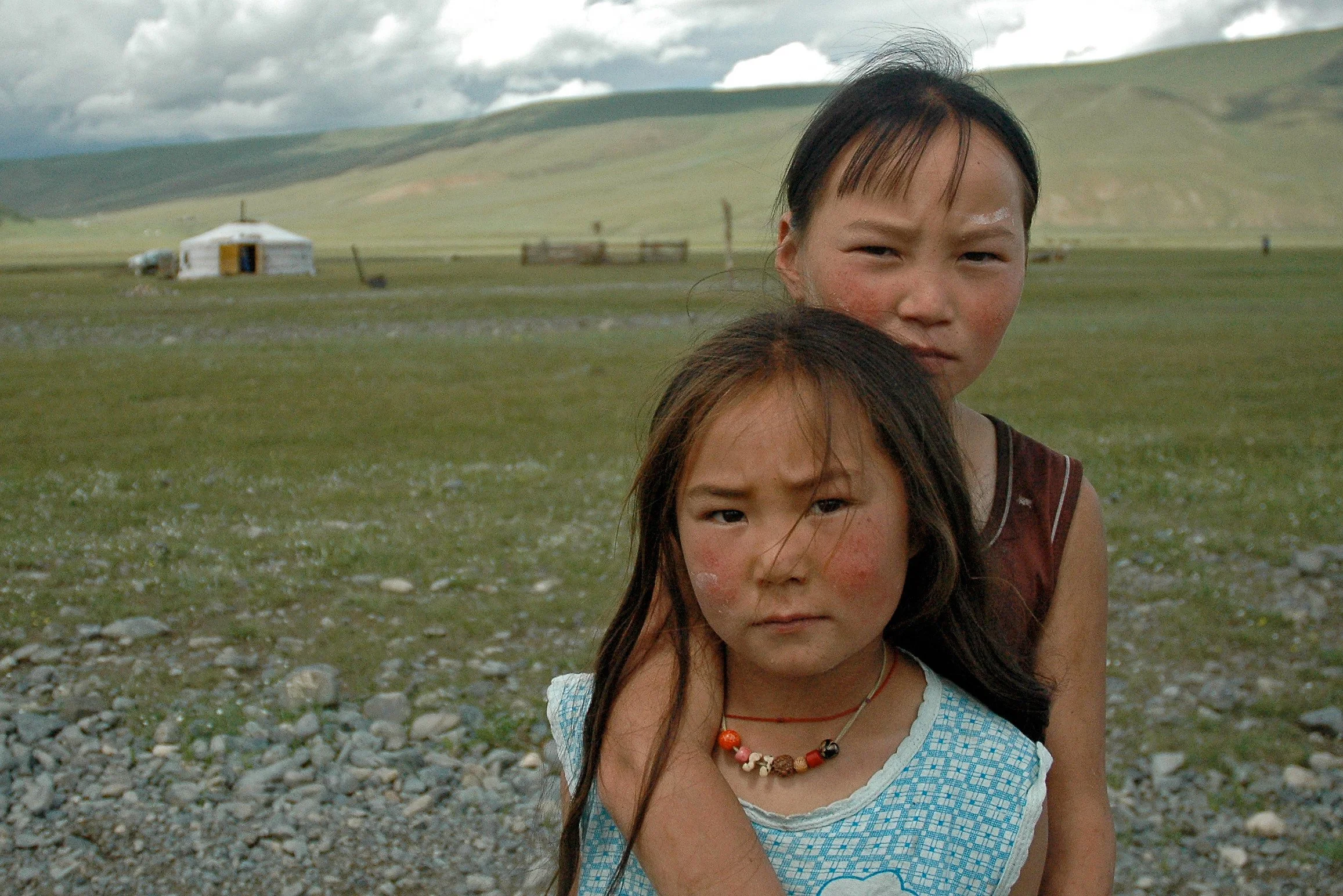 Two young girls with prominent cheekbones and traditional necklaces standing outdoors in a grassy landscape with a yurts and mountains in the background.