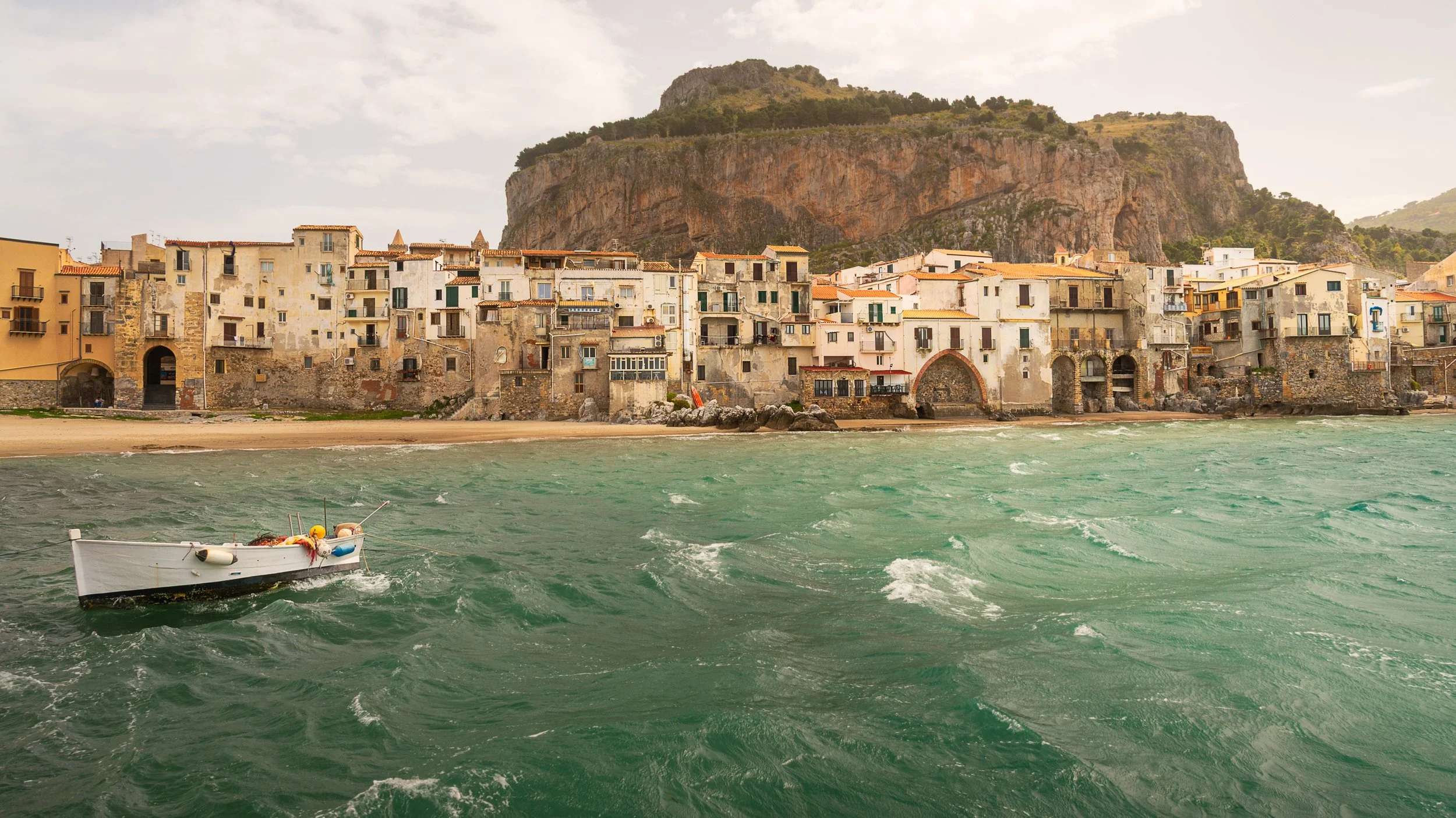 A seaside view of an old town with colorful buildings on the shore and a rocky hill in the background, with a small boat in choppy green water in the foreground.
