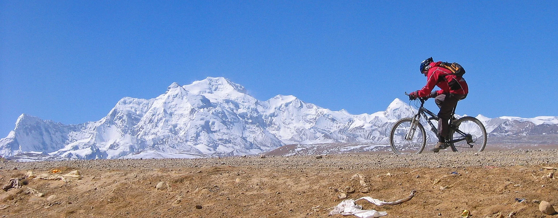 A person wearing a red jacket and helmet riding a mountain bike on a dirt trail with snow-capped mountains in the background under a clear blue sky.