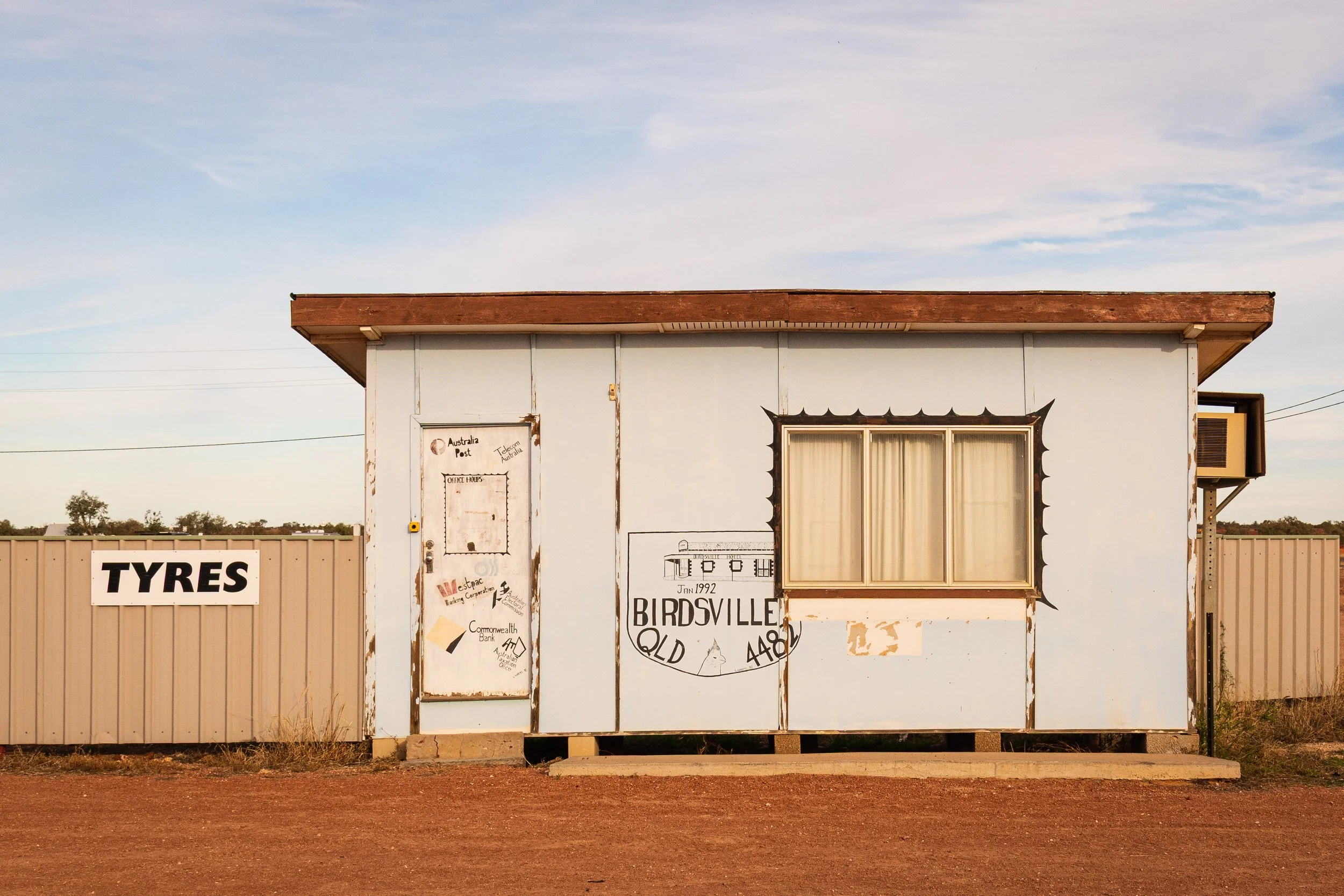 Small, weathered light blue building with graffiti and signs, including a large 'TYRES' sign on beige fence, dirt ground, and cloudy sky.