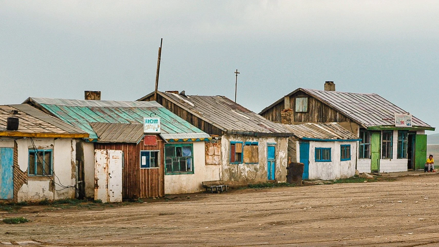 A row of run-down, weathered houses with rusty metal roofs on a dirt ground, with a woman sitting on the far right side.