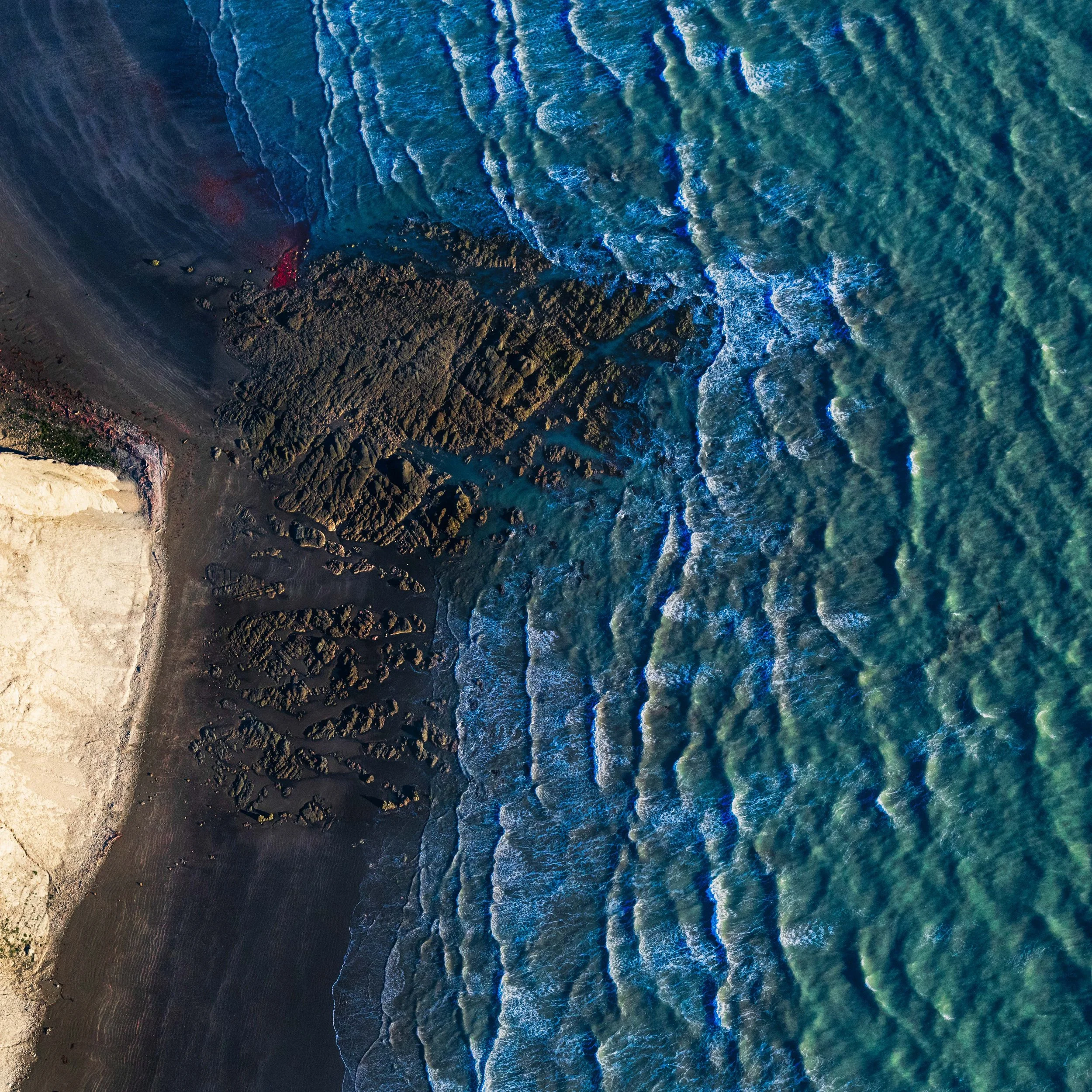 Aerial view of a rocky coastline with waves crashing onto the shore and a sandy beach on the left.