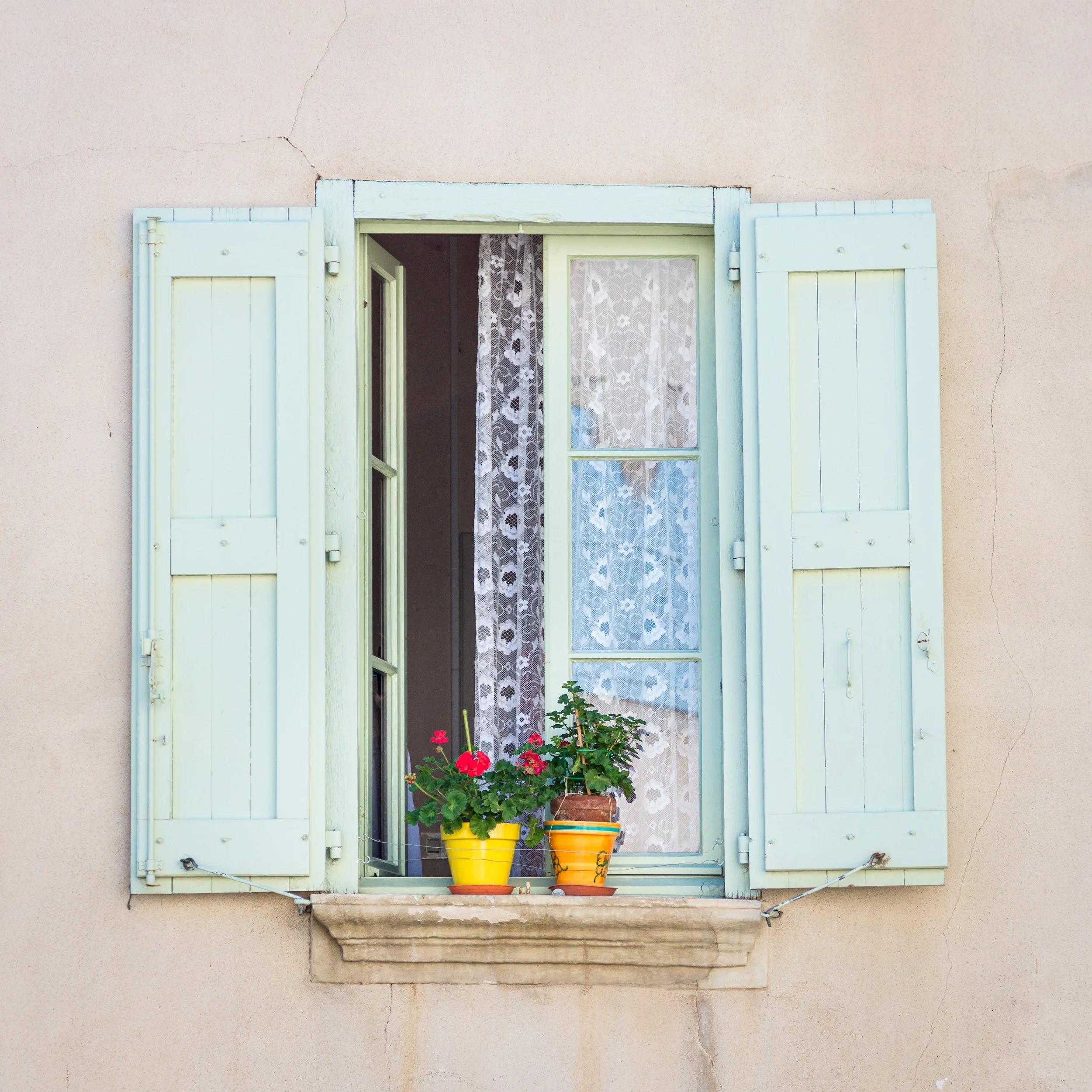 Open window with light blue shutters, lace curtains, and two flower pots on the windowsill with red and pink flowers on a beige wall.