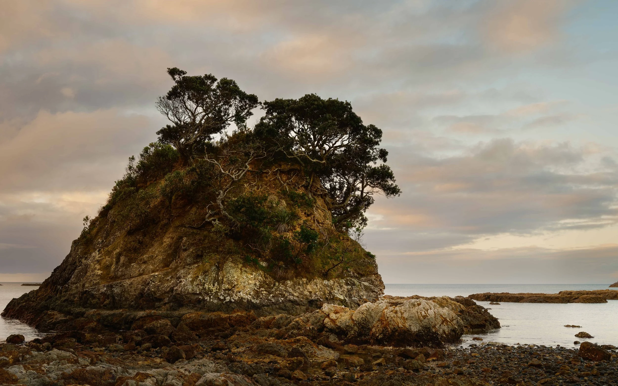 A large rocky island with trees on top, surrounded by smaller rocks and calm water, under a cloudy sky during sunset.