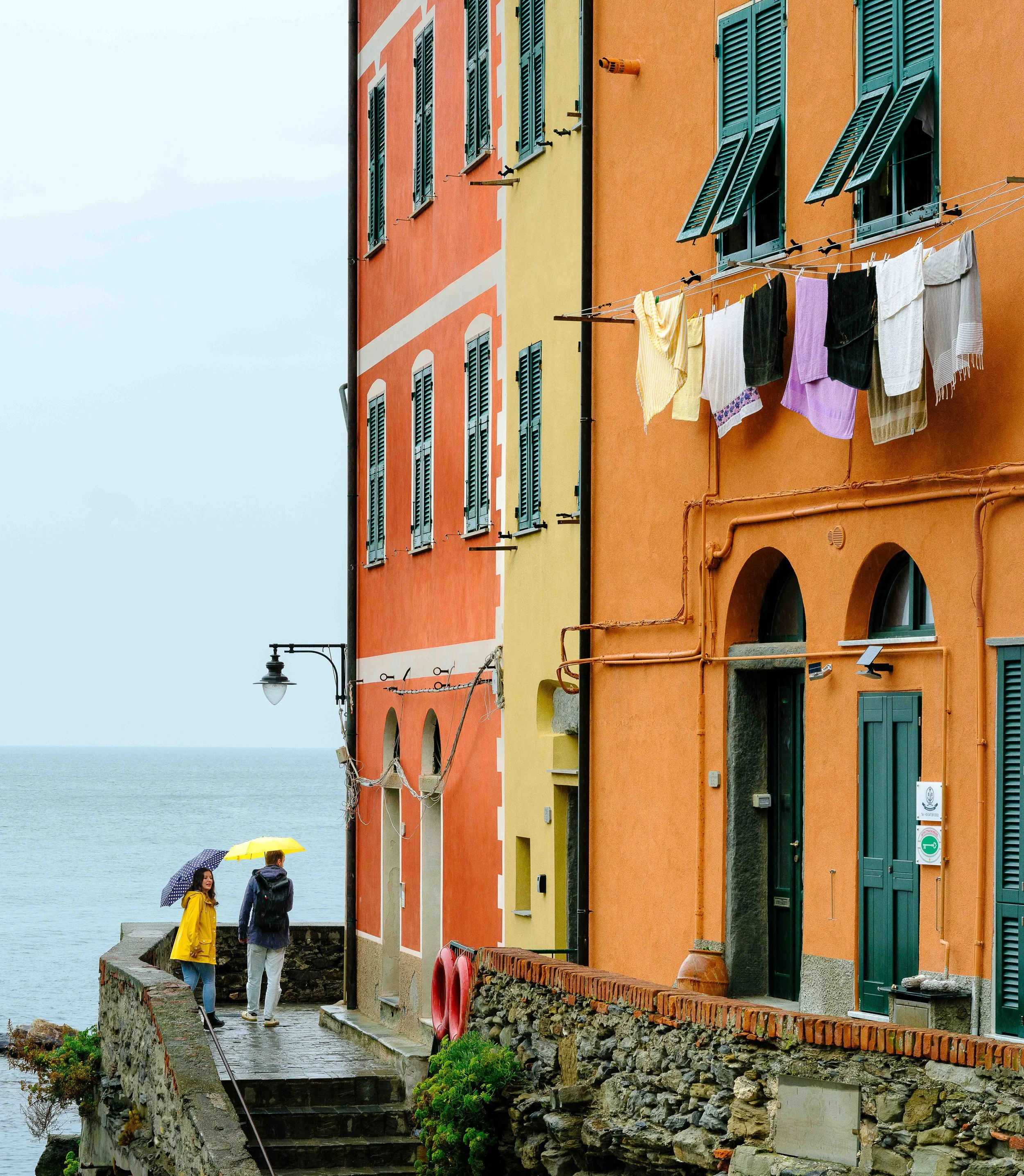 Colorful buildings along a seaside street with laundry hanging on a line and two people walking under umbrellas on a rainy day.