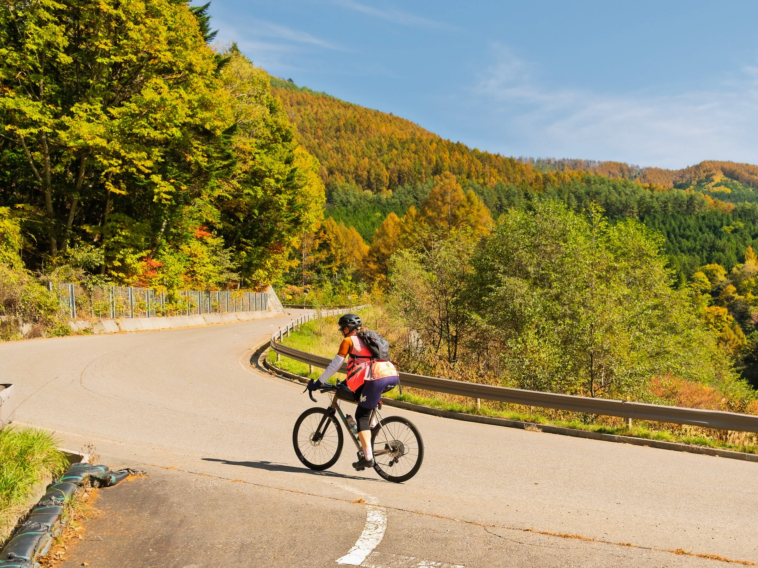 A cyclist rides up a winding mountain road surrounded by colorful autumn trees and hills under a blue sky.