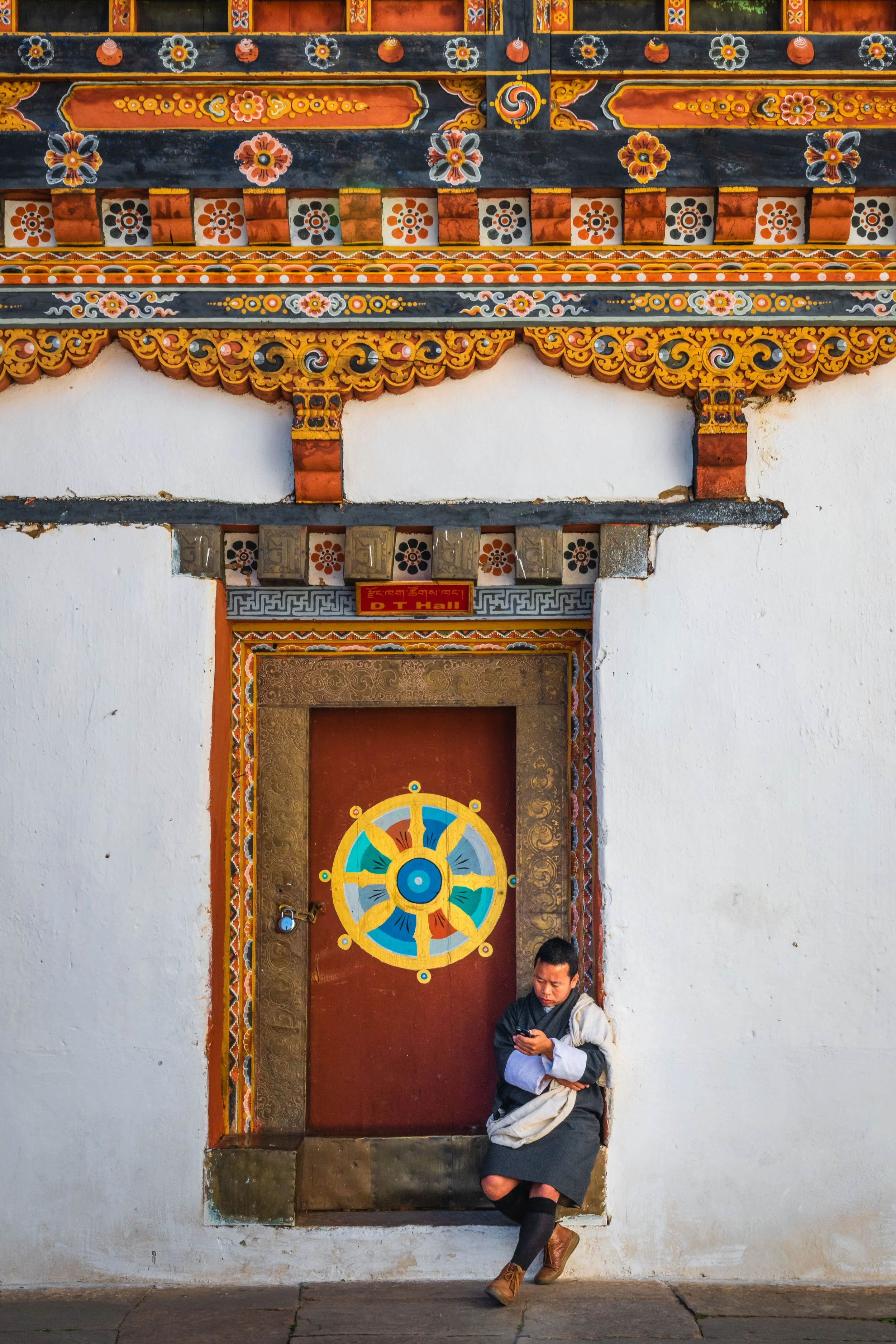 A person leaning against a decorative wall with colorful traditional Tibetan architectural details, including a painted door with a wheel symbol, and sitting on the sidewalk, absorbed in their phone.